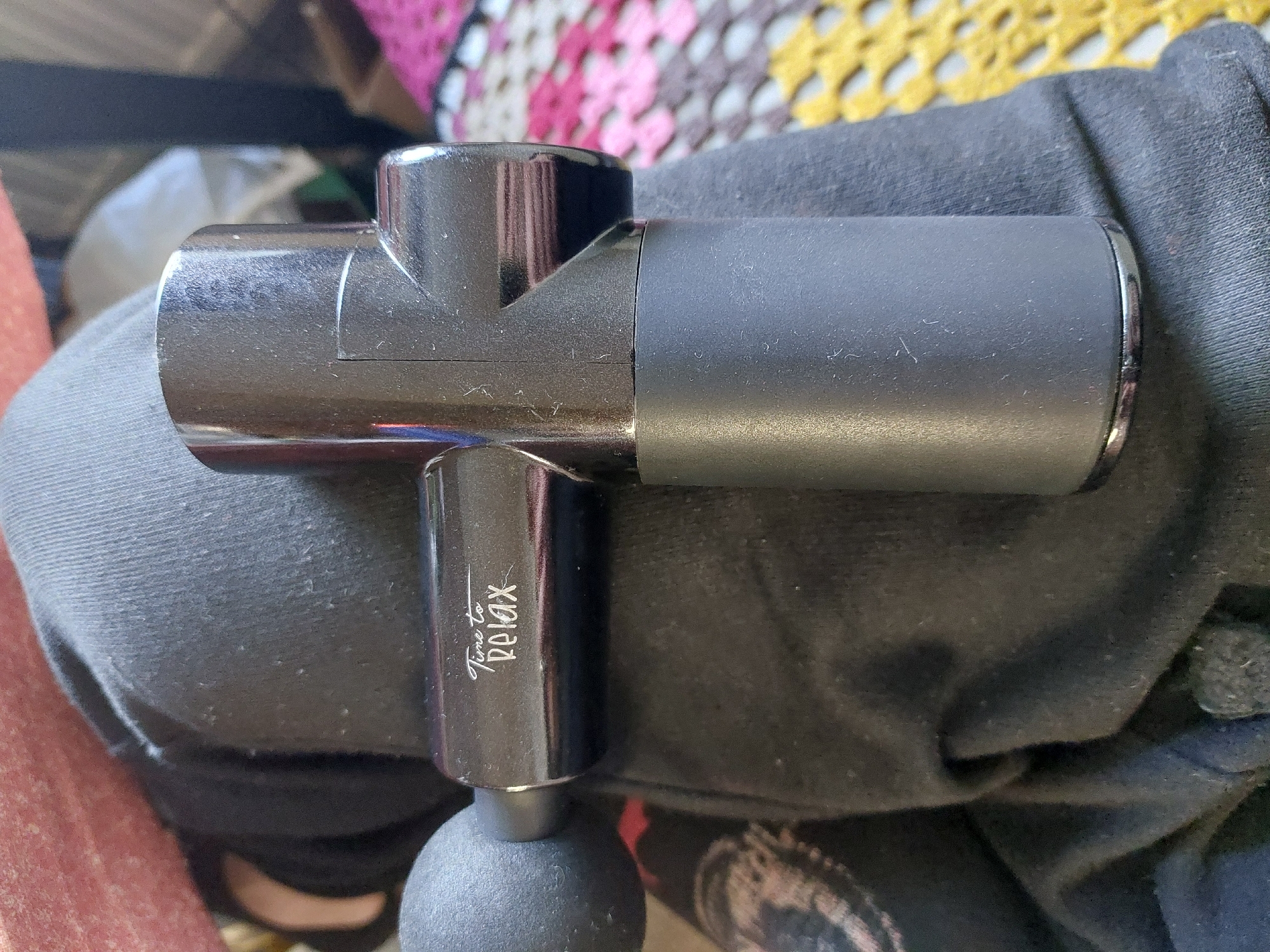 The image shows a black massage gun, labeled "Time to Relax," resting on a dark gray fabric surface, possibly clothing. In the background is a blurred, multicolored knit blanket or rug. The setting appears to be indoors, and there's no information to conclude this image was taken in Marchtrenk, Austria.
