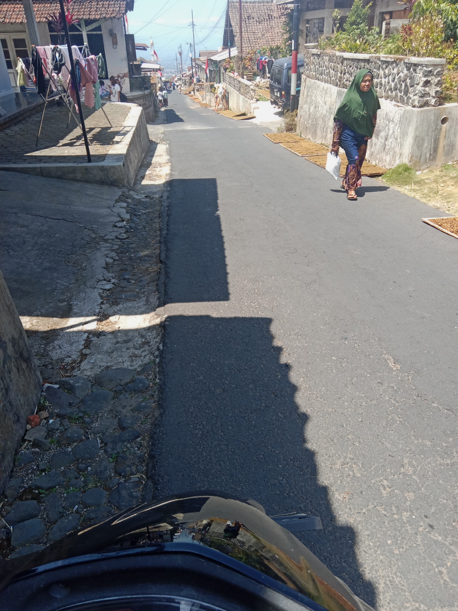 A medium shot captures a sun-drenched, paved road in Mranggen Tengah, Indonesia, viewed from the perspective of a vehicle, likely a motorcycle, due to the handlebars visible at the bottom of the frame. A woman, wearing a green hijab and a patterned dress with blue jeans and sandals, walks uphill towards the camera, carrying a white plastic bag in her right hand. She is on the right side of the road, passing by a low stone wall and some dried goods laid out on mats. To the left of the road, a steep embankment is visible with a concrete structure and a clothesline laden with laundry. In the background, houses with tiled roofs line the street, and utility poles with wires crisscross the sky. The overall scene depicts a quiet, residential street bathed in bright sunlight, casting long shadows across the asphalt.