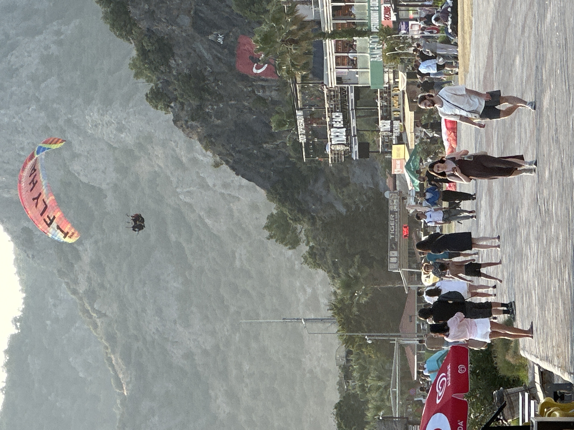The image shows a street scene in Fethiye, Turkey. A mountainside rises to the left of the street. A paraglider is visible in the sky. A crowd of people is walking along the boardwalk. There are buildings, signs, and advertisements visible in the background. A JAK sign is on the mountainside.