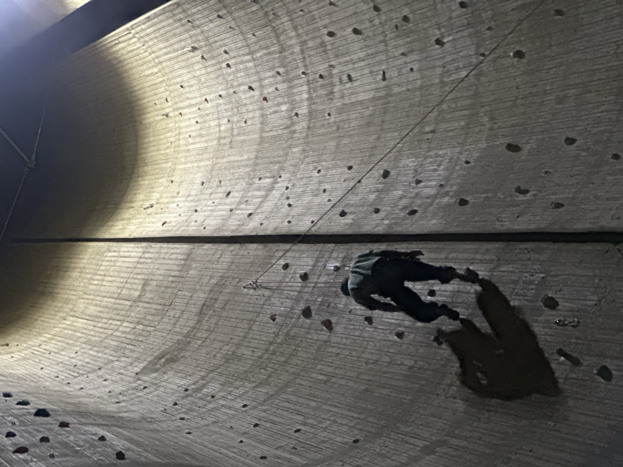 The image depicts a person rock climbing on a curved, man-made climbing wall indoors, possibly in a training facility or recreational center. The wall is studded with climbing holds of various shapes and sizes. A climbing rope is visible, attached to the climber and presumably to an anchor point above. The climber is wearing dark clothing and is positioned at an angle on the wall. A dark shadow of the climber is cast onto the wall beside the person. The location in the city of Unknown, Unknown is unknown.