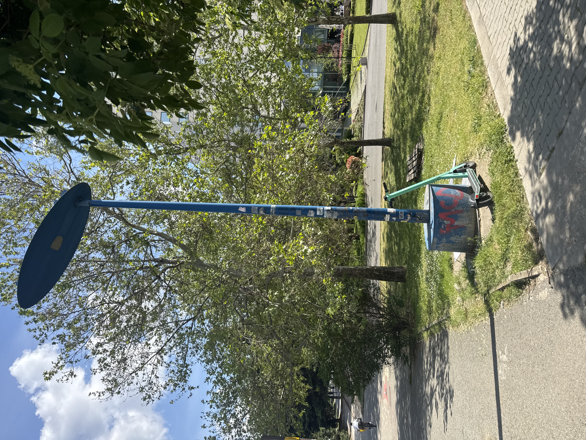 Here's a forensic description of the image:
**Location:** The image appears to be taken on a street in Sofia, Bulgaria.
**Objects:**
* A tall, blue structure with a circular/oval element at the top is present. The pole has visible stickers.
* A blue trash receptacle stands adjacent to the pole.
* A green kick scooter is leaning against the trash receptacle.
* A paved walkway runs along the right side of the frame.
* Lush green grass borders the walkway on one side.
* Trees with green foliage are visible, providing shade.
* A modern building with glass windows is visible in the background.
**People:**
* A single person is riding a bicycle far in the back of the scene.
**Environment:**
* The scene is well-lit, suggesting daytime.
* The environment is urban.