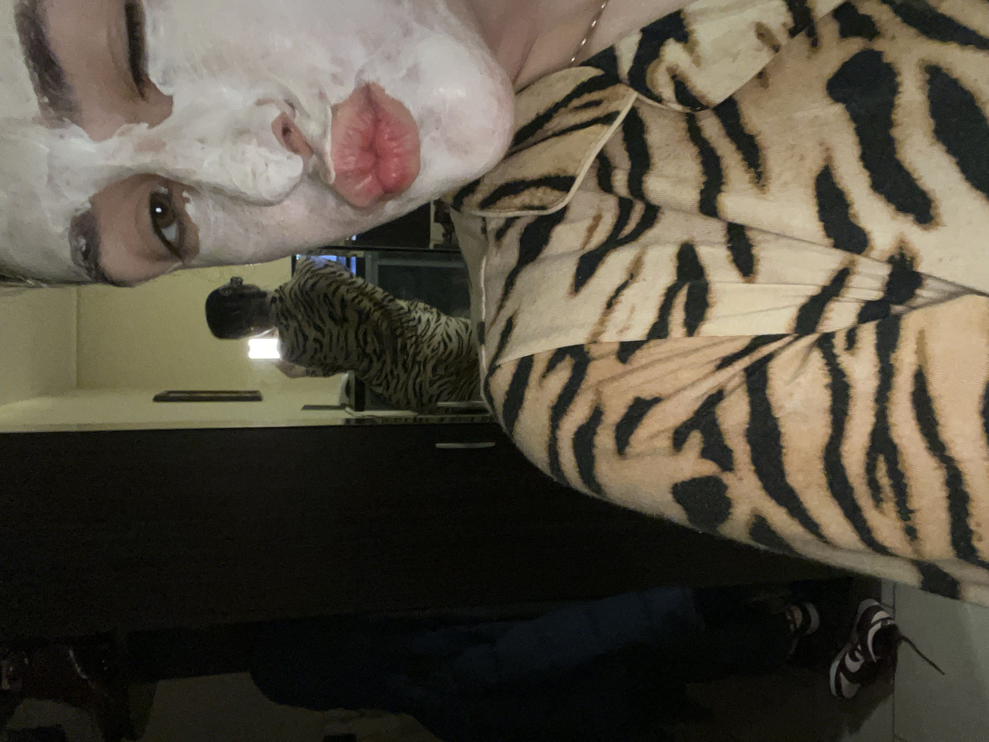 A selfie of a person wearing a tiger-striped pajama top with a white facial mask applied to their face. The person's face is positioned in the upper left of the frame. Only one eye is fully visible. The rest of the face is covered by the white mask. The pajama top dominates the right side of the image. The person is reflected in a mirror. Below the mirror, parts of a dark blue pair of pants and one black and white athletic shoe are visible. The location and city are not identifiable in the image.