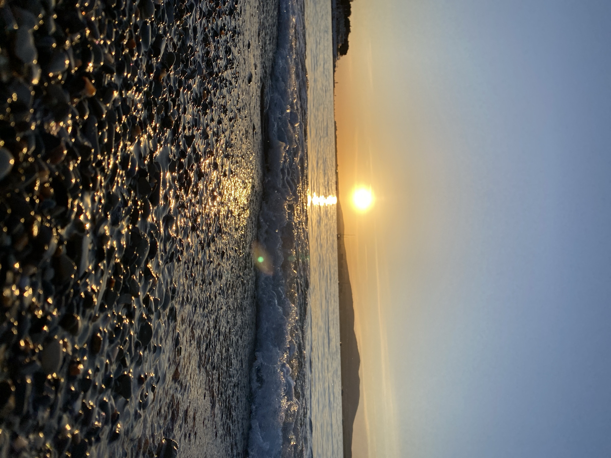 This serene landscape captures a coastal scene during either sunrise or sunset. In the foreground, a pebbled beach is visible, its wet surface glistening with numerous golden reflections from the low-lying sun. Individual dark pebbles are highlighted by bright, sparkling glints of light. A gentle wave breaks onto the shore, forming a line of white foam and creating a dynamic edge between the land and the water.
The sea extends into the midground, reflecting the warm hues of the sky. A prominent, bright sun is positioned on the horizon, casting a radiant glow across the water and into the sky. In the distance, a dark silhouette of land or mountains can be seen along the far horizon line. The sky transitions from a warm, clear yellow-orange near the sun to a soft, pale blue higher up. The overall atmosphere is one of calm and natural beauty, characteristic of a peaceful moment on the coast of Gelendzhik, Russia. There is no visible text in the image.
