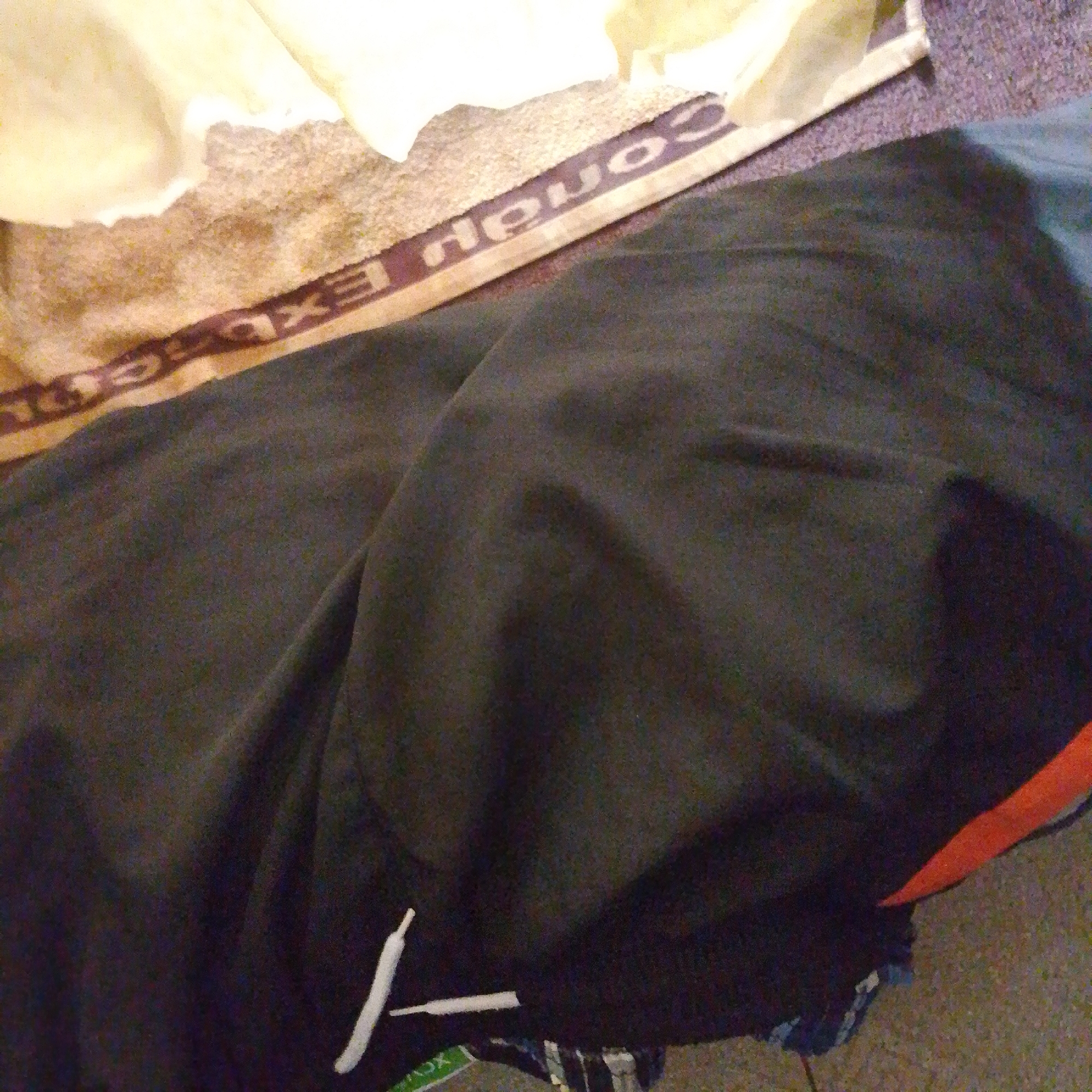 The image depicts a close-up, overhead view of dark fabric, likely clothing, draped over a textured surface. A band with embossed lettering, possibly a brand name or logo, runs across the upper portion of the image. The fabric exhibits folds and creases, suggesting it is soft and somewhat pliable. In the lower right corner, a strip of bright orange material is visible, contrasting with the dark fabric. The overall lighting suggests an indoor setting with a possible light source from above. There are no discernible elements in the image that would indicate its location in Jalingo, Nigeria.