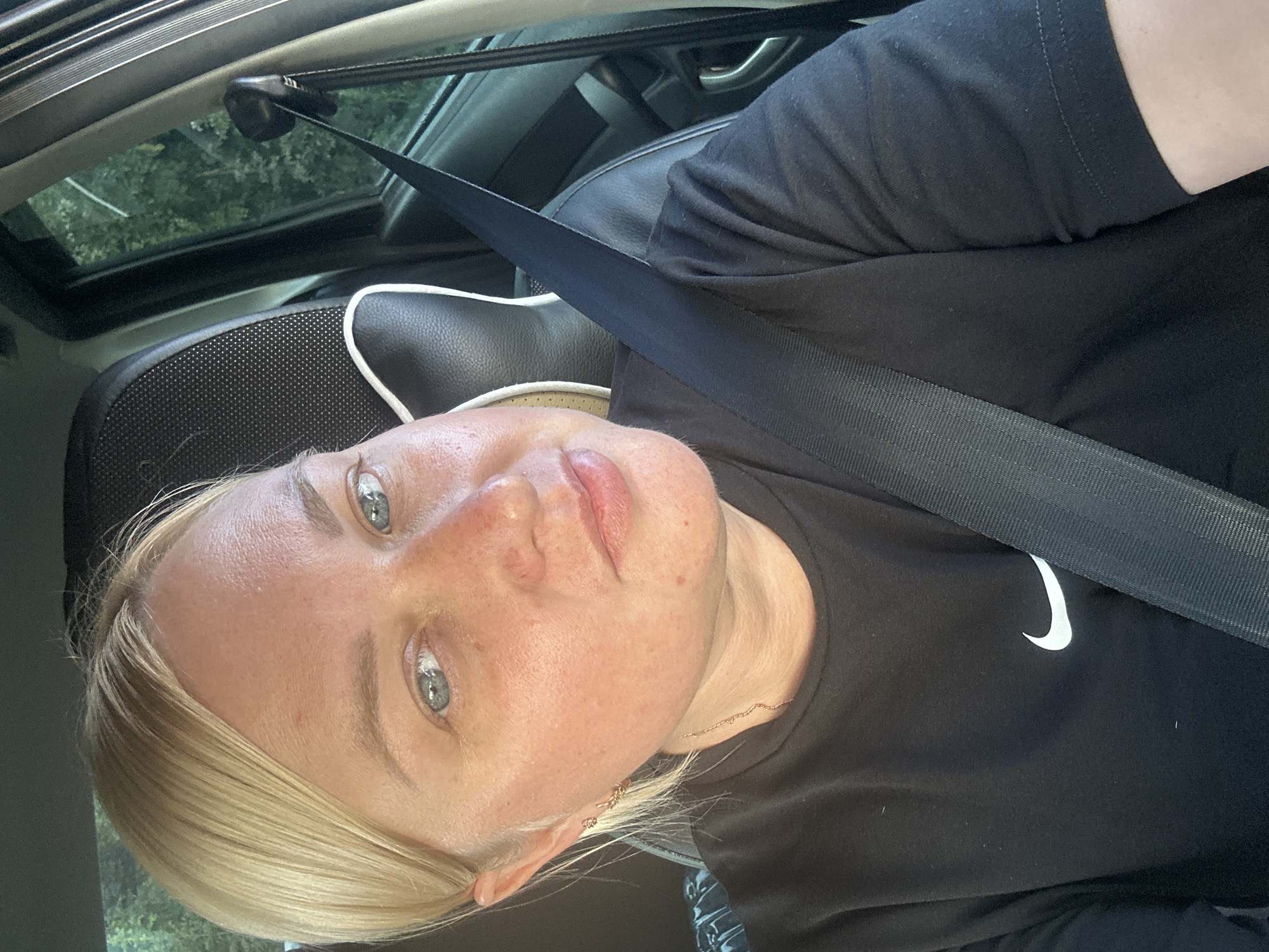 A blonde woman with light blue eyes is sitting in the driver's seat of a car, wearing a black t-shirt with a white Nike logo. A black seatbelt is diagonally across her chest. The background shows the car's interior, including a headrest and a window with trees visible outside. The image appears to be a selfie taken in Chaikovsky, Russia.