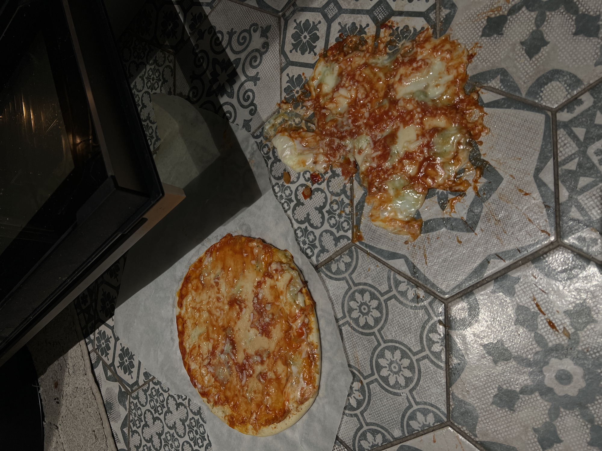 In a domestic setting in Khimki, Russia, a kitchen floor with hexagonal grey and white patterned tiles is depicted. On the left, an unbaked or partially baked pizza, approximately 20-25 cm in diameter, rests on a sheet of white parchment paper. It is topped with red tomato sauce and melted white cheese. To the right, a significant portion of a similar pizza is splattered across the tiled floor, showing a chaotic mixture of red tomato sauce and melted white cheese, indicating it was recently dropped. In the upper right background, a dark-colored oven with a reflective glass door is partially visible, with an internal light source faintly glowing. A light-colored, textured mat or rug is partially visible in the upper left, near the base of the oven. The scene is illuminated by an overhead light source casting shadows. No individuals are present.