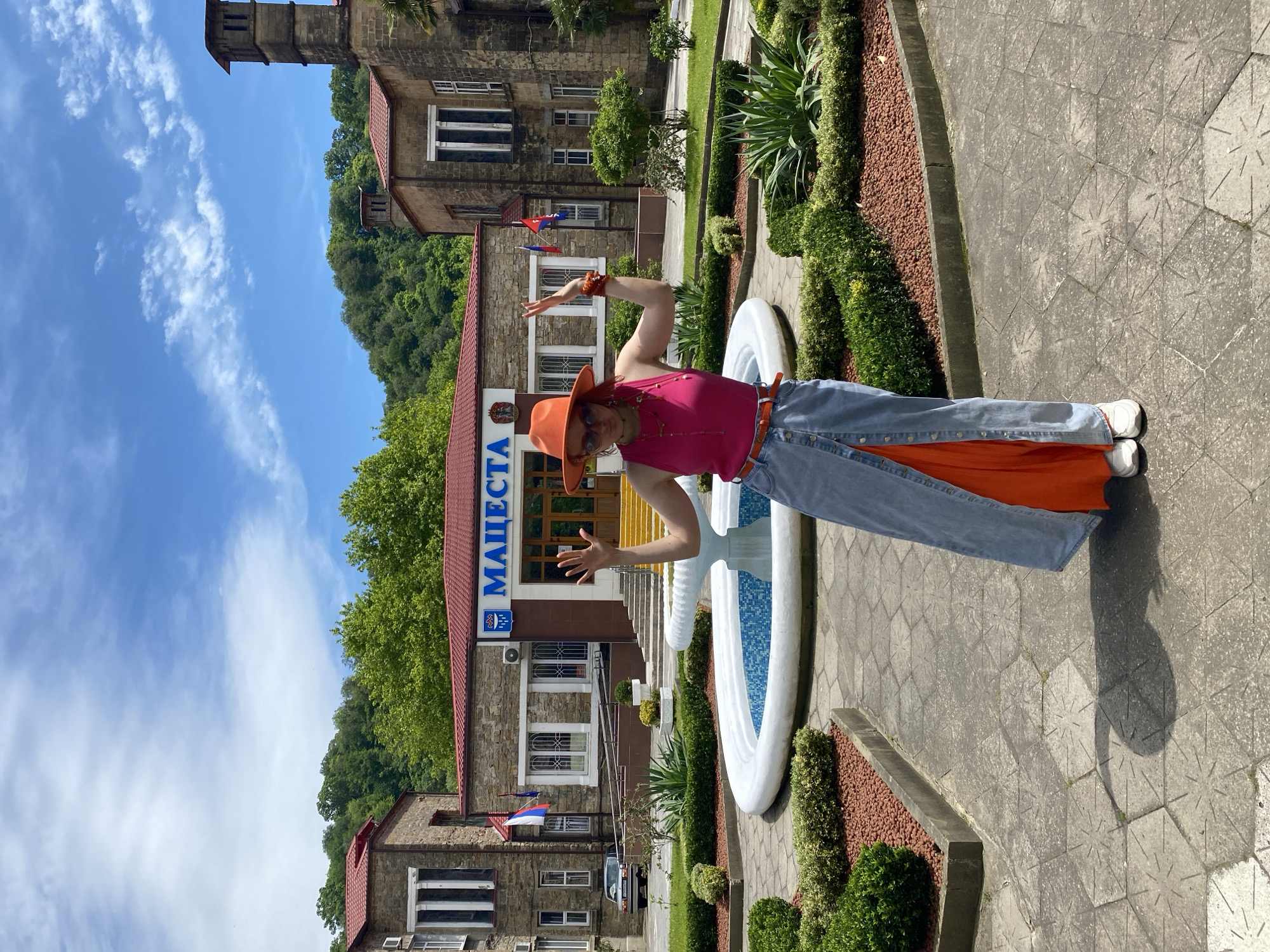 A person stands next to a fountain, with a building bearing the name "МАЦЕСТА" in blue Cyrillic letters in the city of Sochi, Russia. The person is wearing a bright orange hat, a pink tank top, light blue jeans with orange fabric panels peeking from the bottom, a belt, and white sneakers. They have an orange bracelet on their raised left arm. The building in the background is made of stone with a red roof and yellow door frame. To the left of the main building is a tower-like structure. There are Russian flags on the building, and a car is parked in front. The sky is blue with some clouds. The fountain is white with light blue water in the basin. There are manicured bushes and red gravel bordering the fountain. The ground is paved with gray stones. A forested hill is in the distance behind the building.
