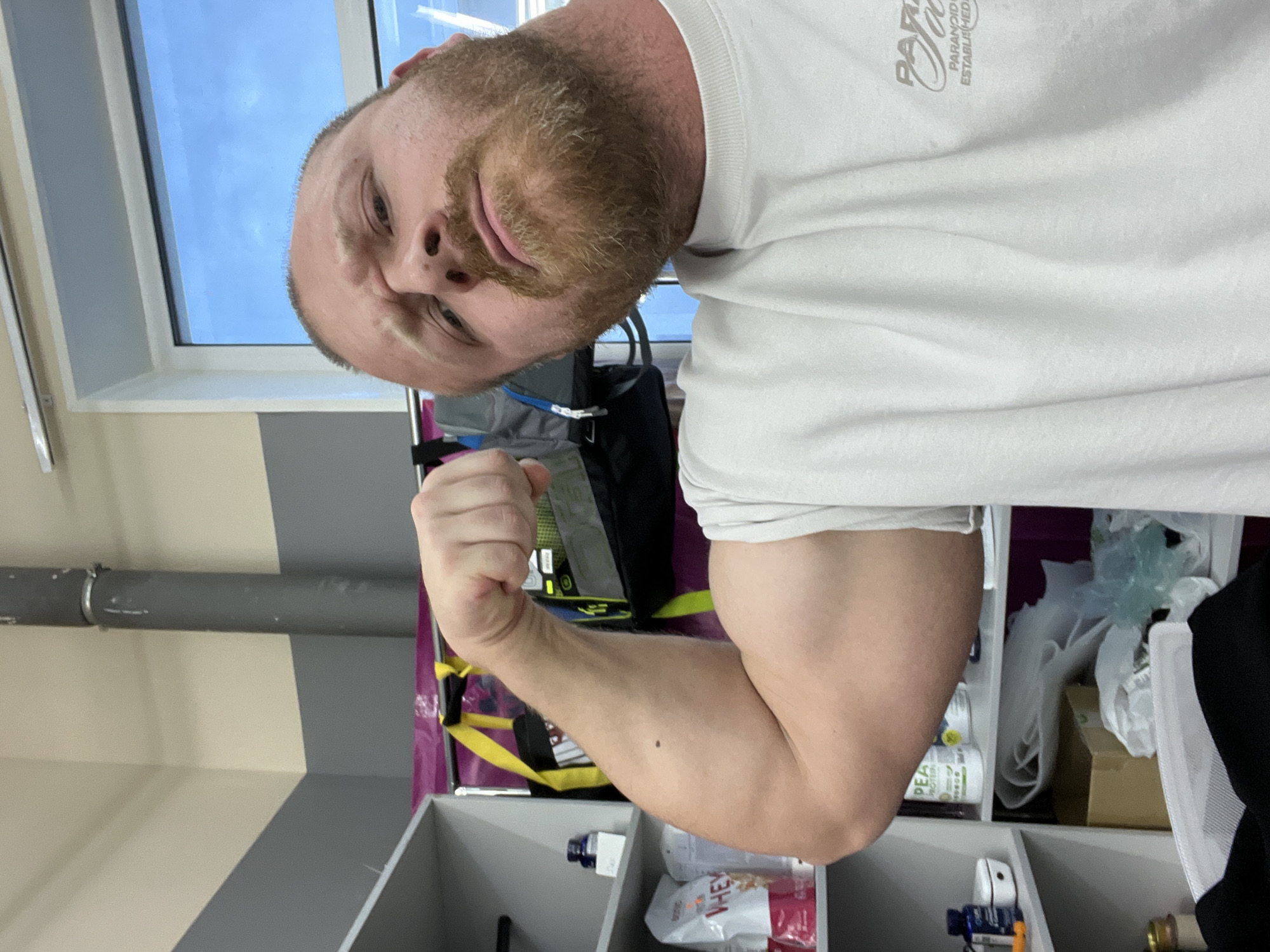 A Caucasian man with a reddish-brown beard and a bald head, wearing a white T-shirt, is flexing his right bicep for the camera. The man is positioned inside a room. Visible in the background are a window with a view of a blue sky, a gray pipe, a gray and beige wall, a black backpack, and shelves with various items including a bag labeled "Whey". The location is specified as Chaikovsky, Russia.