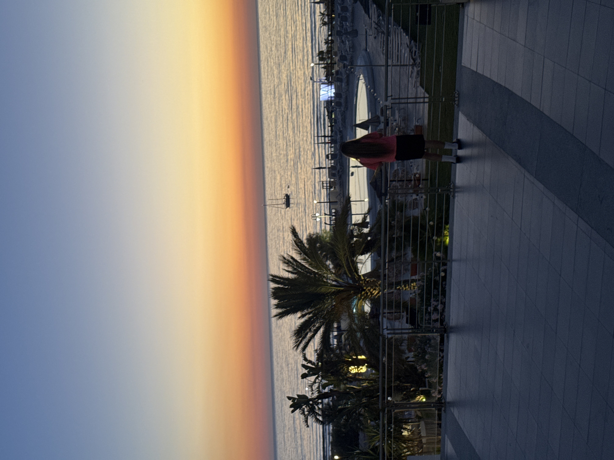 A young person stands on a tiled balcony overlooking a beach resort at dusk. The sky transitions from a pale blue at the top to a warm orange and pink hue near the horizon, indicative of sunset. The sea is calm with gentle waves. In the middle ground, palm trees frame a resort area with a swimming pool, lounge chairs, and umbrellas. A sailboat is visible in the distance on the water. The person is wearing a pink shirt, a dark skirt, and white socks with dark shoes, facing away from the camera and towards the sunset. The balcony features a metal railing. The overall setting suggests a relaxing evening at a coastal location in Isola delle Femmine, Italy.