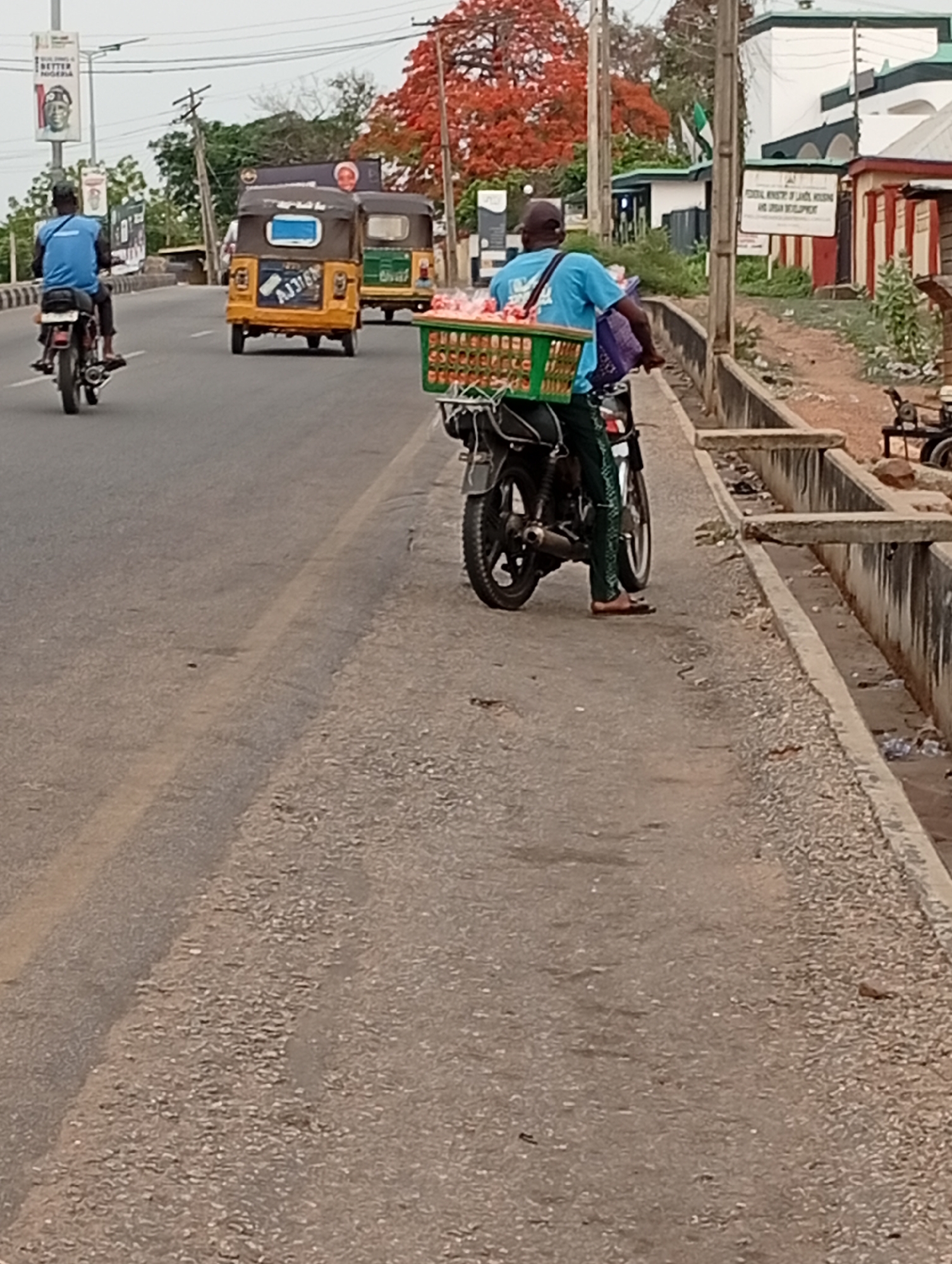 The image shows a road scene in Lokoja, Nigeria. A man in a blue shirt and black cap is riding a motorcycle loaded with a green basket filled with produce. He is on the right side of the road, close to a concrete drainage ditch. The drainage ditch is visible, and there is litter inside. Another motorcycle is further ahead on the road to the left, along with two yellow auto rickshaws. A sign is visible with the words "FEDERAL MINISTRY OF LAND, HOUSING, AND URBAN DEVELOPMENT". The trees behind the man have orange flowers. Banners are visible in the distance along the road. The road is cracked and uneven in places.