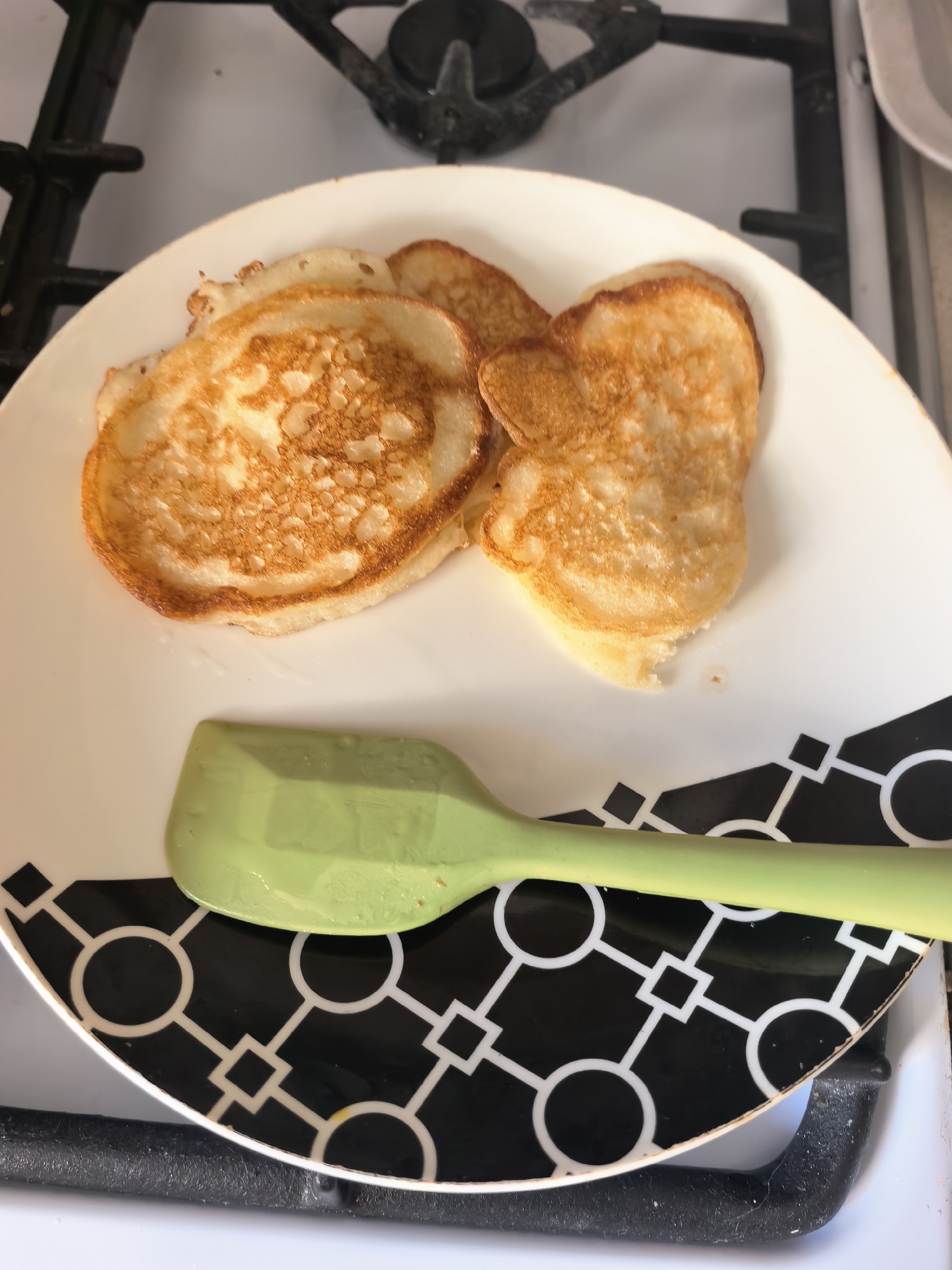 A plate with three pancakes and a green spatula sits on a stovetop. The plate is white with a black and white geometric pattern along the edge. The pancakes are golden brown with textured surfaces. The stovetop is white with black burner grates. The image was taken indoors. There is no information available to determine the location within the city of Sterlitamak, Russia.