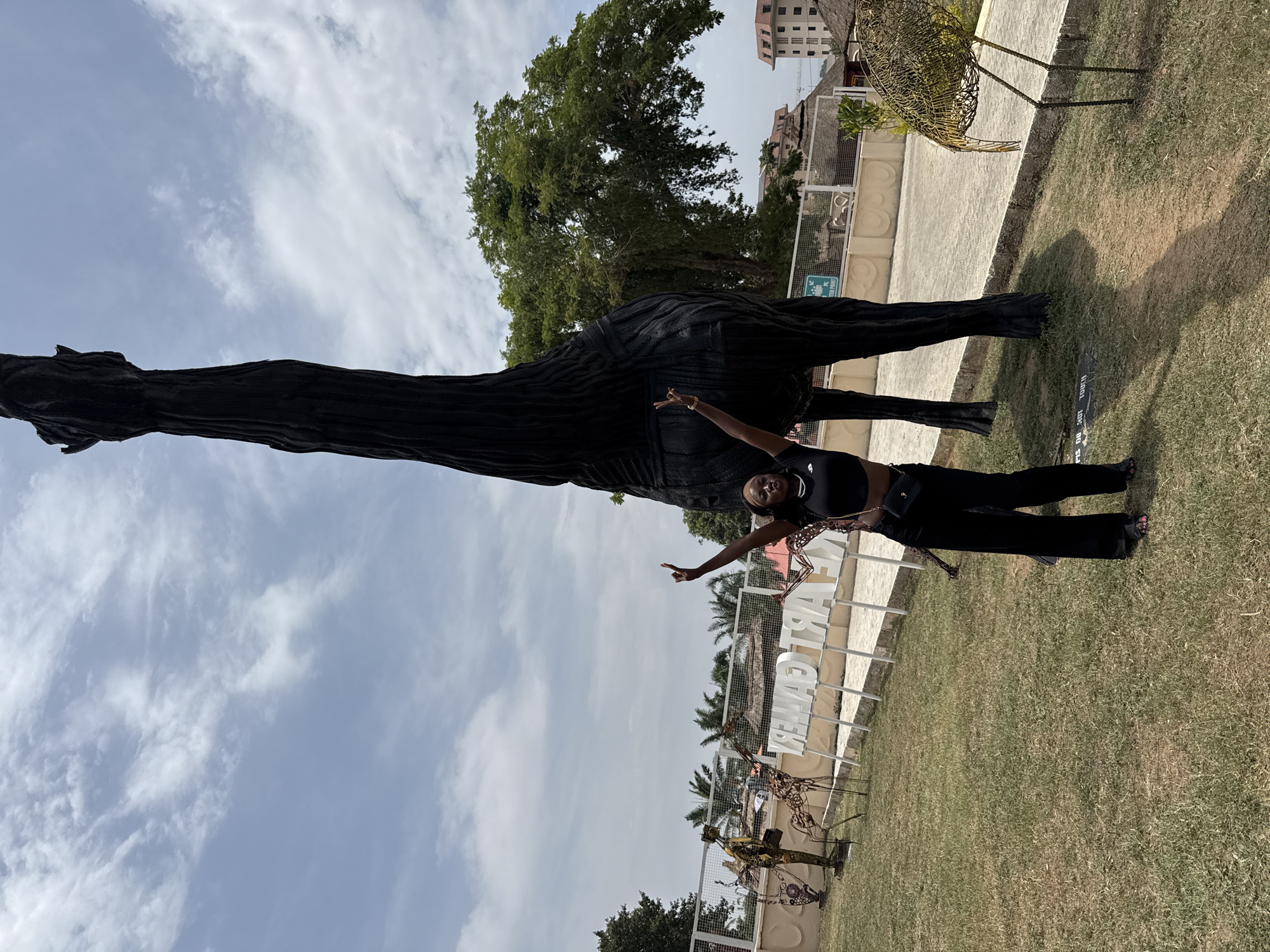 The image shows an outdoor scene at the Mamy Art Gallery in Abuja, Nigeria, captured during bright daylight under a partly cloudy sky. In the foreground, a woman stands smiling, with her arms outstretched and hands forming peace signs. She is dressed in a black sleeveless top, black trousers, and sandals, and appears to be posing playfully for the camera.
Dominating the left side of the frame is a massive, dark-colored sculpture of a giraffe, its textured surface suggesting it might be made from recycled materials. Its long neck extends high into the sky. To the right of the woman, a prominent white sign on the grassy lawn clearly spells out "MAMY ART GALLERY" in large block letters. Nearby, several smaller, intricate metal sculptures, resembling a bird and possibly other animals, are also visible on the grass.
The scene features a well-maintained grassy area, concrete pathways, and mature green trees in the mid-ground and background. Multi-story buildings can be seen in the distance behind the trees. A smaller, dark sign on the grass near the base of the large giraffe sculpture reads "PLS DO NOT TOUCH." The overall atmosphere is bright and inviting, reflecting a pleasant day for visiting an art gallery.