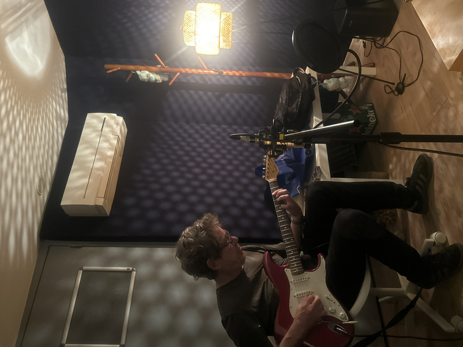 An adult male, appearing middle-aged with light brown, wavy hair and glasses, is seated on a white office chair, actively playing a red electric guitar in an interior room designated as Paris, France. He wears a dark short-sleeved shirt, dark trousers, and dark athletic shoes (Adidas Gazelle-style). His left hand is fretting the guitar neck, and his right hand is positioned over the bridge pickup area, engaged in strumming or picking.
The room features dark blue/purple sound-dampening wall panels with a visible grid pattern. A lighter grey/white wall section or door is visible to the left, with a blank rectangular frame mounted on it. An off-white air conditioning unit is mounted high on the dark wall. Distinctive dappled light patterns from a textured source are cast upon the ceiling and upper walls.
A multi-tiered, woven or rattan-style floor lamp emits warm light in the right corner, adjacent to a dark wooden coat rack holding two small, light-colored plush figures. A white rectangular table is positioned behind the guitarist, holding a blue fabric bag, a black backpack, and other small objects. A black microphone on a stand, equipped with a circular pop filter, is positioned between the guitarist and the table. A small black rectangular speaker or amplifier unit rests on the light wooden floor to the right of the microphone stand. The overall scene suggests a personal music studio or rehearsal space.