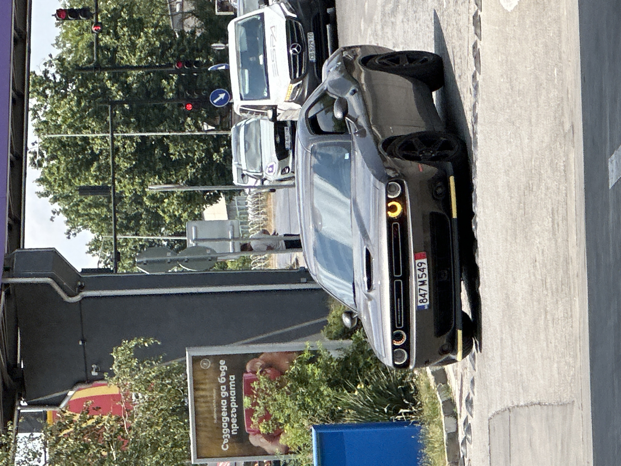 In Veliko Tarnovo, Bulgaria, a dark gray Dodge Challenger with license plate 847 M549 is positioned on a paved road, facing forward and slightly to the right. The car has yellow accents on its front bumper and black rims. Behind the Challenger, a white Mercedes-Benz van, marked with "ESPA" and a license plate that appears to be CD 8229 SO, is also visible. Further back, a silver Ford SUV with license plate CB 4670 BH and a white van are present.
The scene is outdoors, with trees and foliage contributing to the environment. A large advertisement billboard is situated to the left of the Challenger, featuring text in Bulgarian. Traffic lights are visible in the background. A person is standing near the billboard, and another is near the traffic lights. The road surface shows markings and speed bumps.