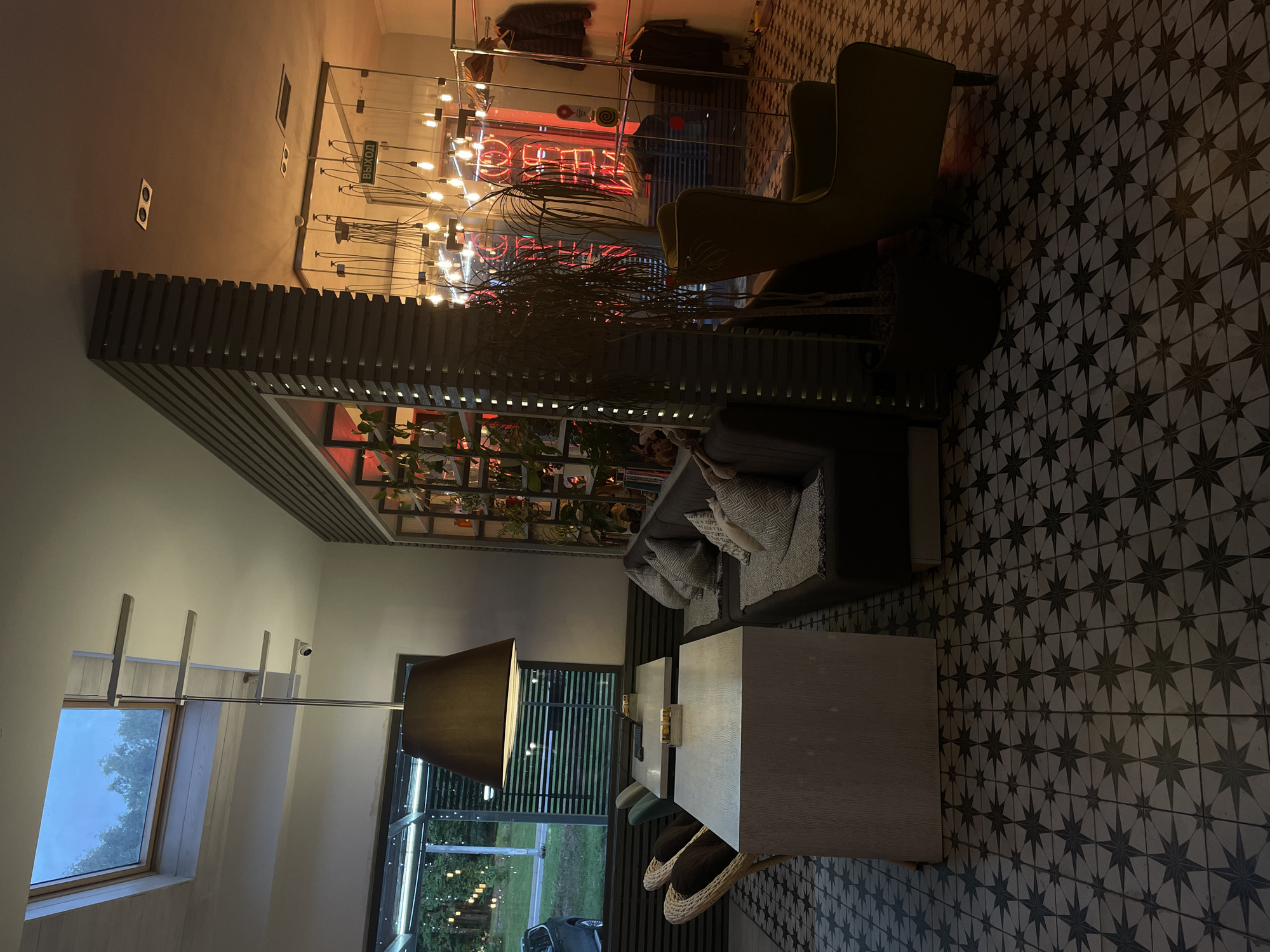 This media file shows an indoor scene, likely a cafe or restaurant, with a mix of modern and vintage elements. The setting appears to be dimly lit, suggesting it might be evening or night.
In the foreground, a large, dark lampshade hangs over a table, casting a warm glow. The table itself is a light-colored, rectangular surface, with some yellow items, possibly small bottles or containers, placed on it. Two wicker chairs with dark cushions are tucked under the table. Further back, a grey sofa with patterned cushions is visible, along with a section of a stylish wooden divider.
The background reveals more of the establishment. There's a wall with exposed light bulbs and a neon sign, possibly in red, that is partially obscured. A sign with Russian text, "ВЫХОД" (Exit), is also visible. A green armchair is positioned near a glass partition, behind which more lighting and some items are arranged. The floor is tiled with a decorative pattern of stars.
A large window on the left side of the image looks out onto a treelined area, with a dark sky visible, reinforcing the idea of it being dark outside. Another window on the right shows a view of string lights in an outdoor area, possibly a patio or garden. A security camera is mounted on the wall above the left window.
No people are visible in the scene, and the overall atmosphere seems relaxed and inviting, with a touch of retro charm due to the lighting and decor. The presence of plants on shelves in the background adds a touch of nature to the indoor environment. The location is identified as Krasnogorsk, Russia.