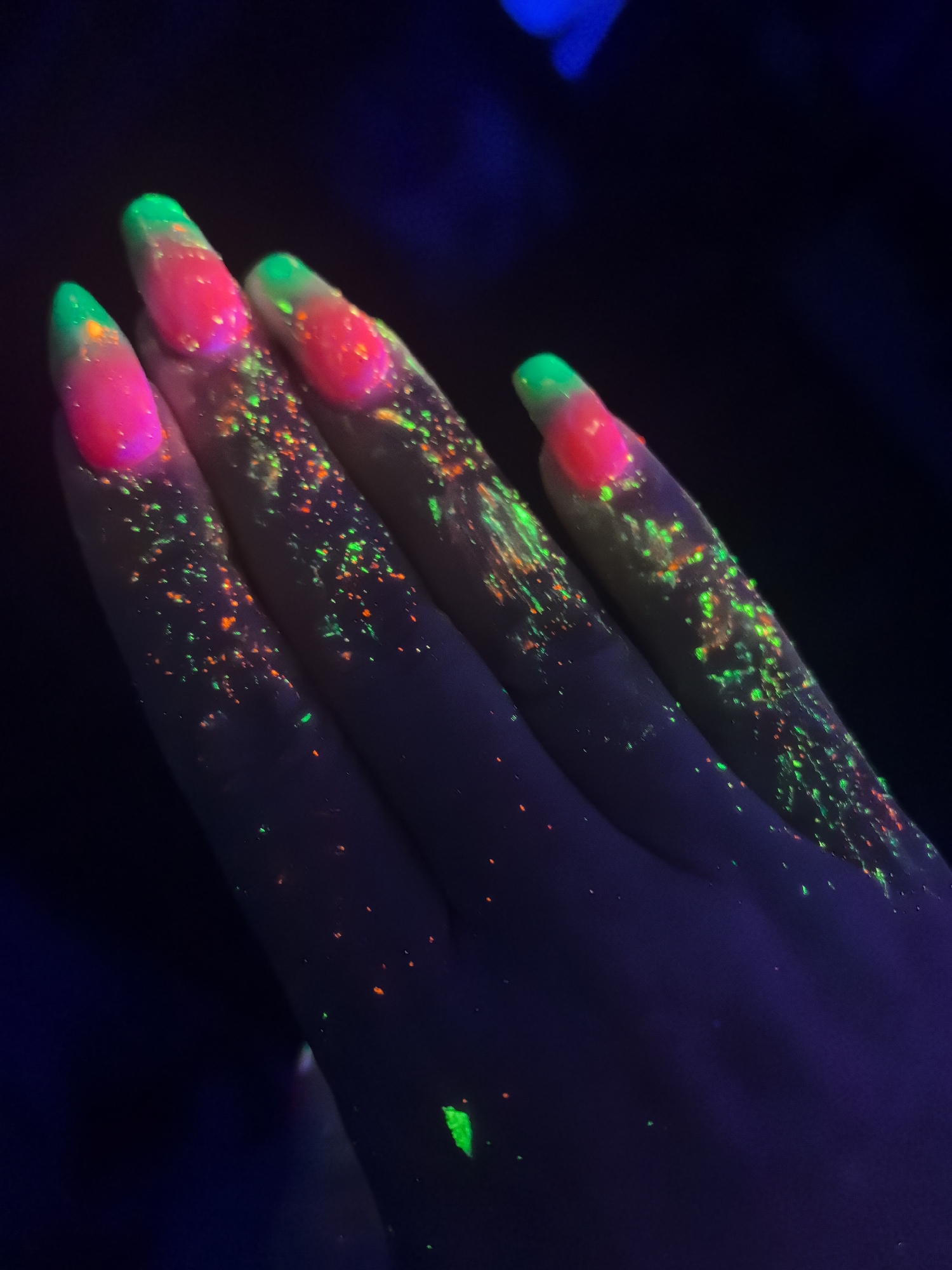 A close-up shot shows a hand under ultraviolet light. The nails are painted with a two-tone design, featuring a pink base and a green tip. The skin and nails have numerous small speckles of green and orange that fluoresce under the UV light. The background is dark, with a slight hint of blue illumination in the upper portion. There is no indication that this image was taken in Noviy, Russia.
