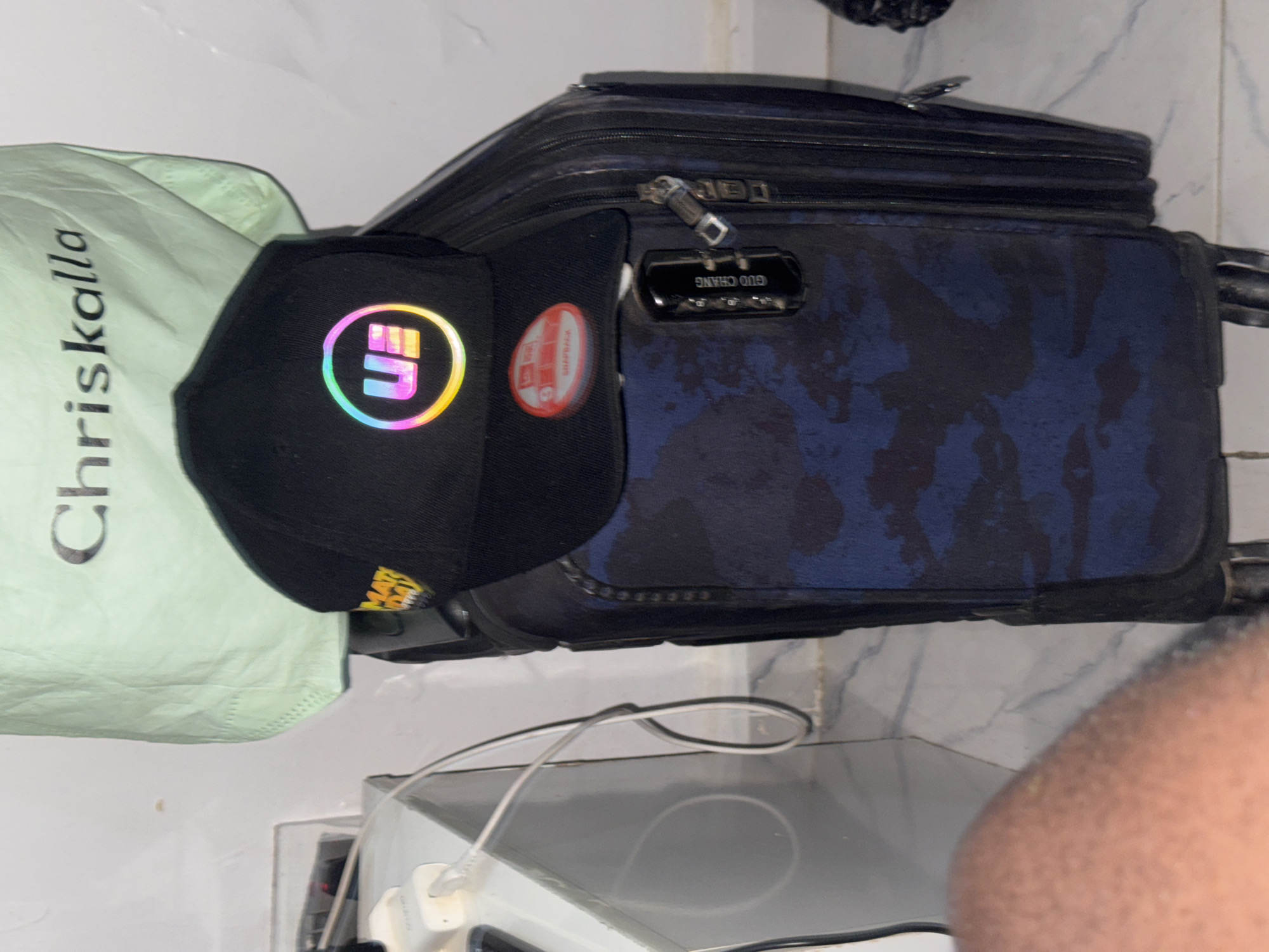 A suitcase with a combination lock sits on a white surface. A black baseball cap with a colorful logo sits atop the suitcase, partially covering a green bag with white lettering. A white charging cable is visible on the surface below the suitcase. The location is Ado, Nigeria.