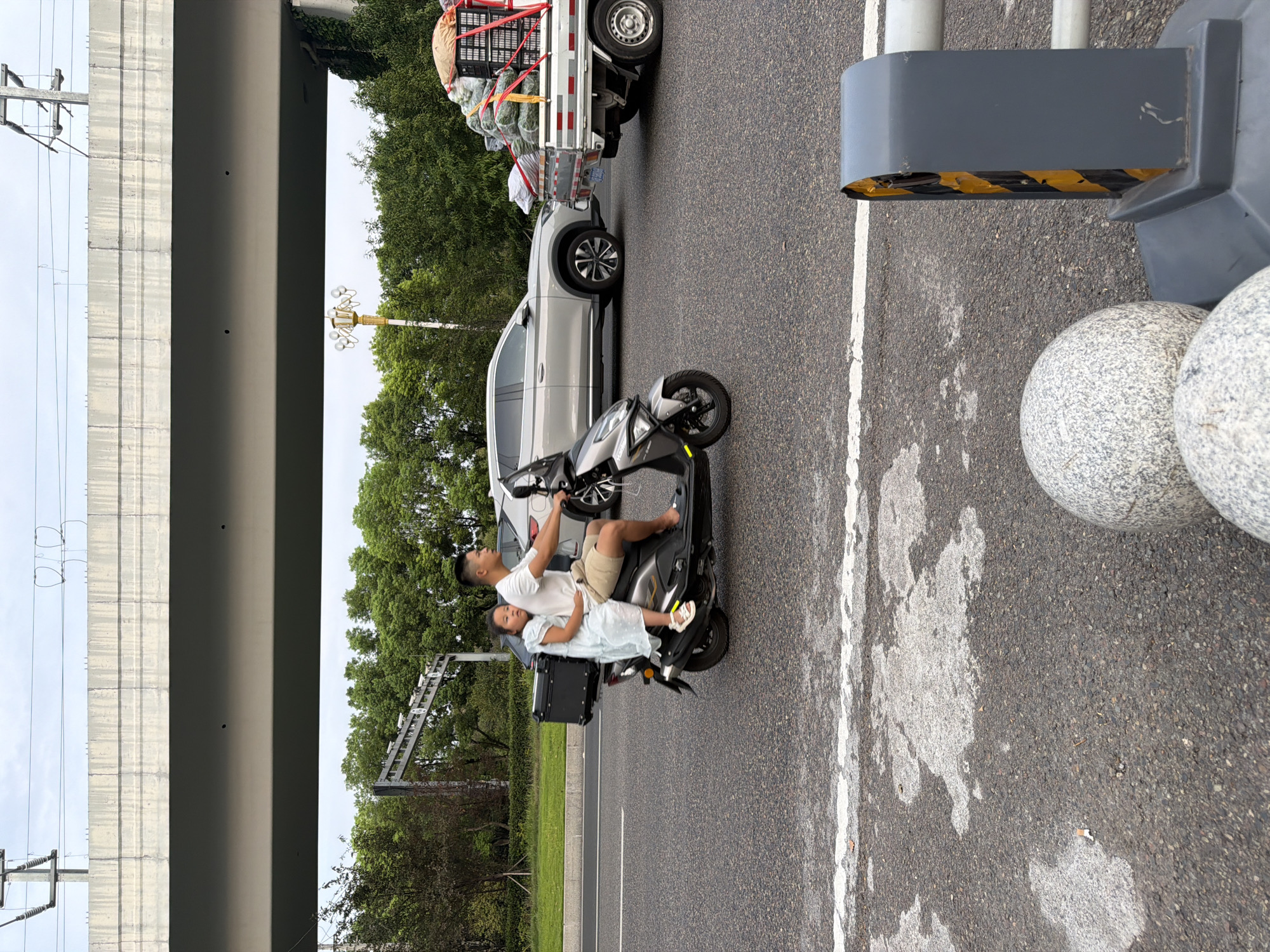 In Jiangjin, China, a man rides a scooter on a road, with a young girl sitting in front of him. The man is wearing a white t-shirt and beige shorts, and the girl is in a light blue dress. They are riding alongside a silver SUV and a truck loaded with produce. The scene is under a concrete overpass, with trees and a decorative lamppost in the background. The road is asphalt with white lane markings.