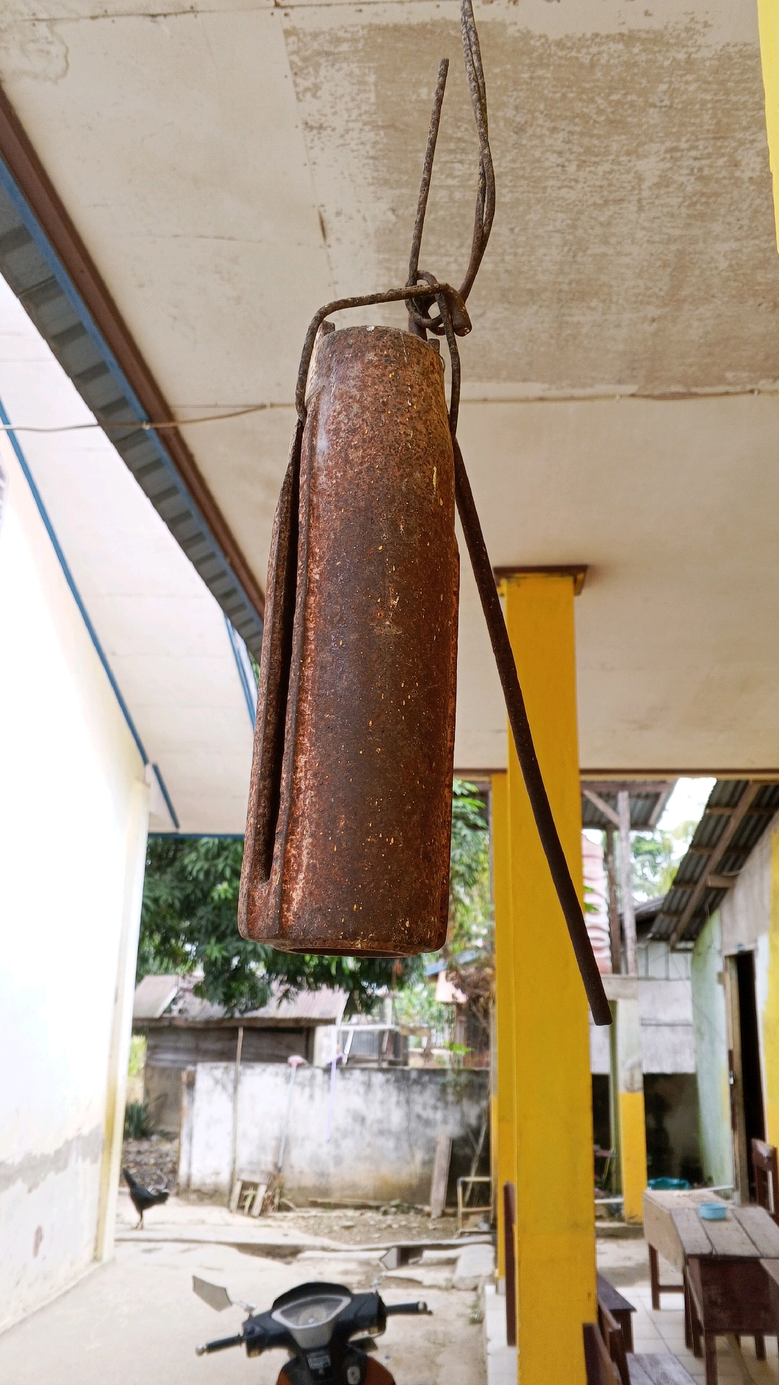 A rusty metal cylinder, possibly a bell or a counterweight, hangs from a wire attached to a structure with a concrete ceiling. The cylinder is roughly cylindrical with a folded section, showing significant signs of rust and wear. A thin, dark rod extends diagonally from the top of the cylinder upwards, also appearing to be metal. The background shows an outdoor scene, likely in a village or rural area, with a yellowish pillar supporting the ceiling structure. To the left, a white building with a blue trim is visible, and further back, there are several smaller structures, trees, and a chicken walking on the ground. In the foreground, the front portion of a black and orange scooter is visible. The overall impression is of an aging, utilitarian structure in a tropical or subtropical environment, possibly in Sritabaang, Indonesia, given the architectural style and vegetation.