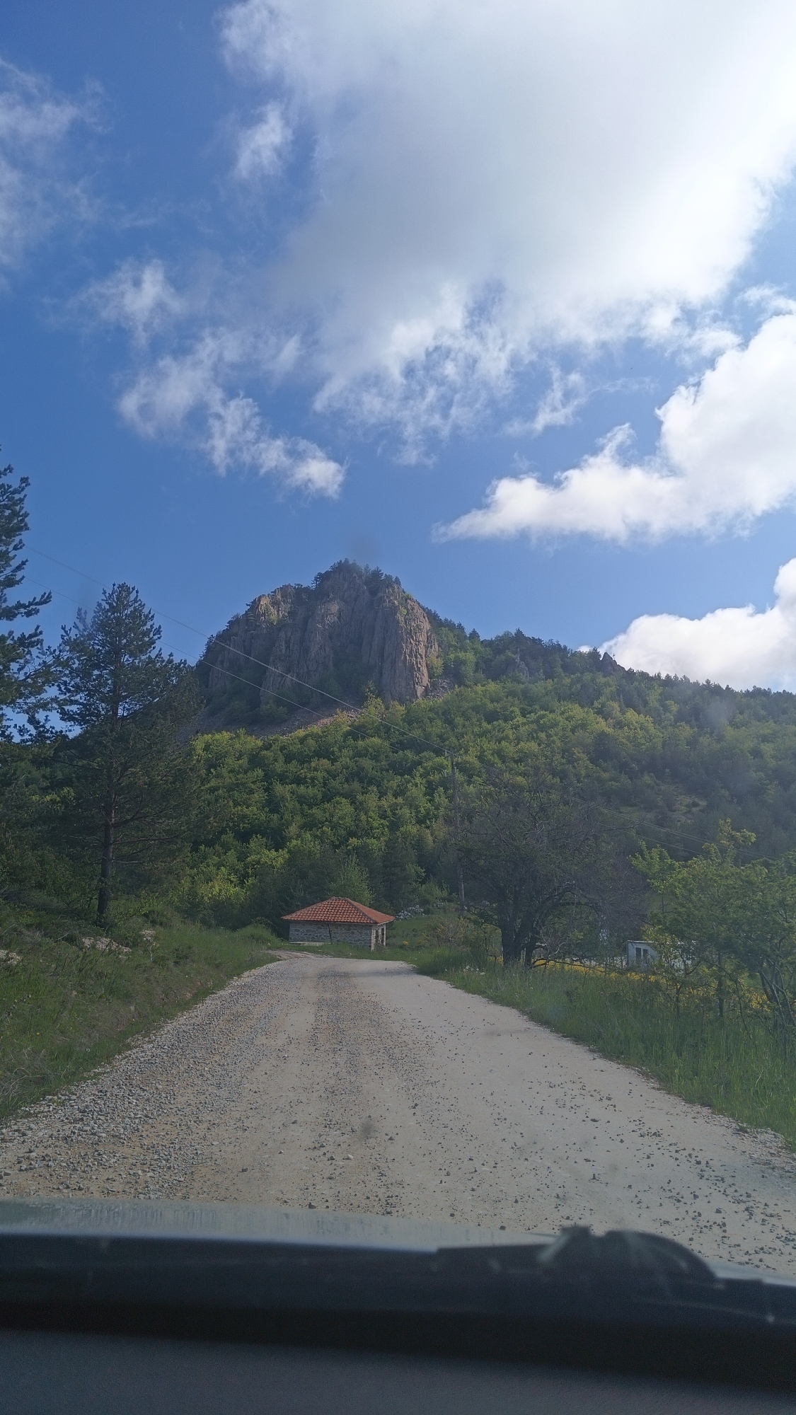 The image depicts a gravel road leading towards a small building with a red tiled roof, set against a backdrop of a mountain covered in trees. The sky above is blue with scattered white clouds. The image is taken from what appears to be inside a vehicle, with a portion of the dashboard visible at the bottom of the frame. This location is within the city of Vrata, Bulgaria.