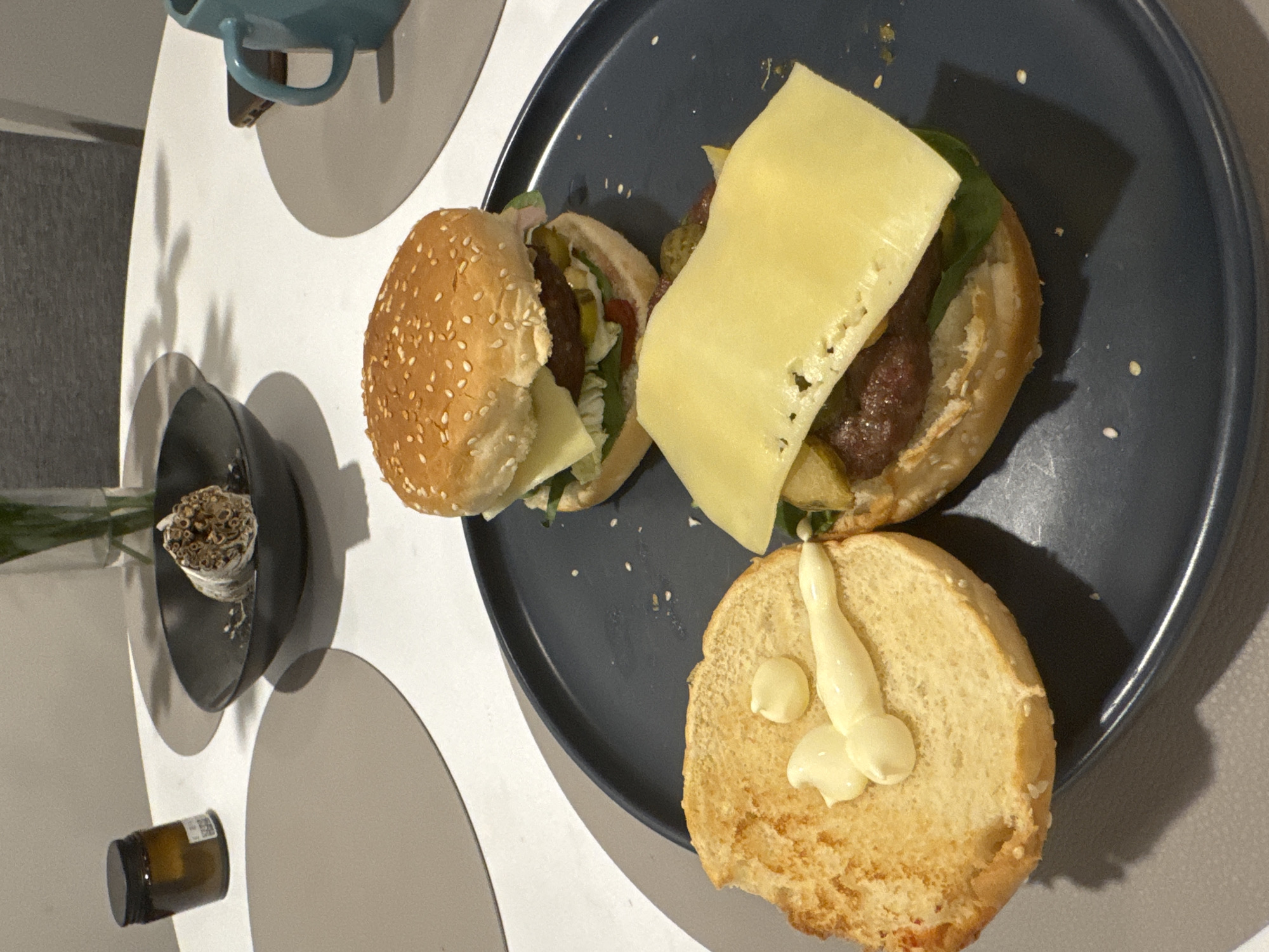 This image captures an indoor tabletop scene, likely in Dubai, United Arab Emirates, focusing on two hamburgers being prepared or served on a dark grey or blue plate. One hamburger is largely assembled, featuring a sesame seed bun, a patty, a slice of cheese, lettuce, and pickles. The other hamburger is partially assembled, with its toasted bottom bun topped with mayonnaise, alongside a patty covered with a draped slice of cheese, lettuce, and pickles. Small white sesame seeds are scattered on the plate.
The plate is situated on a light-colored, possibly white or light grey, table. Distinct shadows cast by the objects suggest indoor lighting. To the left of the main subject, a light blue mug hangs from a fixture. Further left, a small, dark bowl contains a bundle of dried, light-colored cylindrical plant material, possibly incense or dried herbs, positioned next to a glass vase holding a green plant stem. A small, amber-colored jar with a black lid and an illegible label is also visible on the table surface. No people are present, and the time of day and weather are not discernible from the image.
