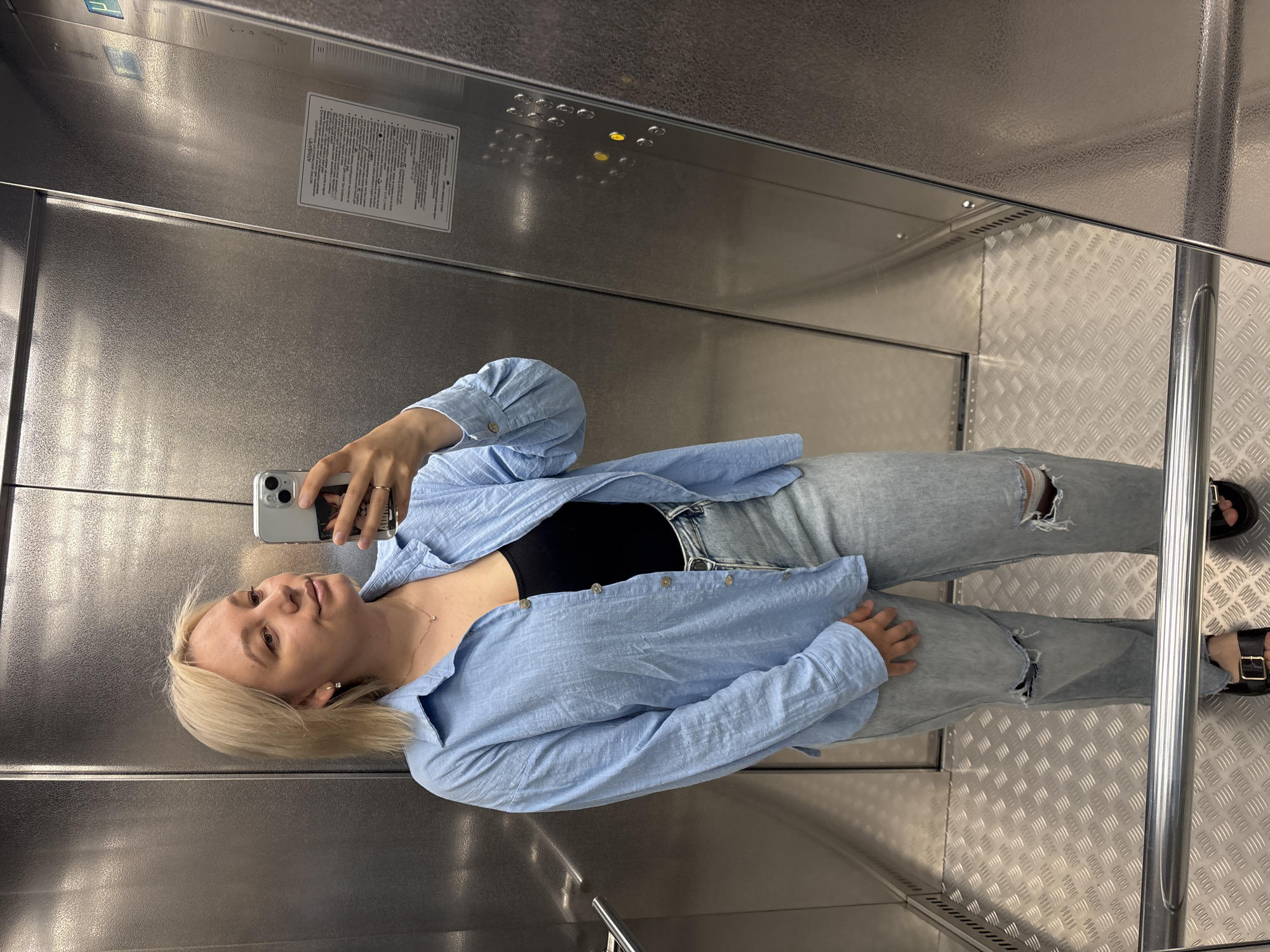 A young woman with blonde hair is taking a mirror selfie in an elevator. She is wearing a light blue, textured, unbuttoned shirt over a black top, and light-wash distressed jeans. She has a delicate necklace with a small pendant and a ring on her right hand. The elevator has stainless steel walls and a diamond plate floor. A warning sign is visible on the right wall of the elevator. The image appears to be taken in Chaikovsky, Russia, as indicated by the text on the warning sign which contains Russian language.