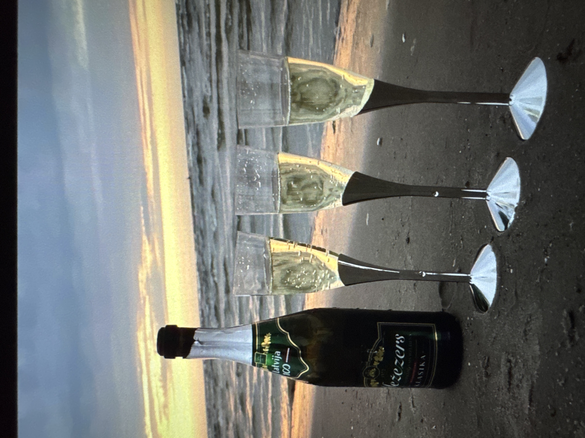 A bottle of champagne and three champagne glasses filled with champagne are on a sandy beach. The bottle has a green and gold label that reads "Latvija 100" and "Jezers". The glasses have clear bowls and silver stems. The ocean is in the background, with gentle waves. The sky is a gradient of blue and orange, suggesting either sunrise or sunset. The overall impression is one of celebration or relaxation at the seaside. It is not possible to determine the specific location within Riga, Latvia from the image.