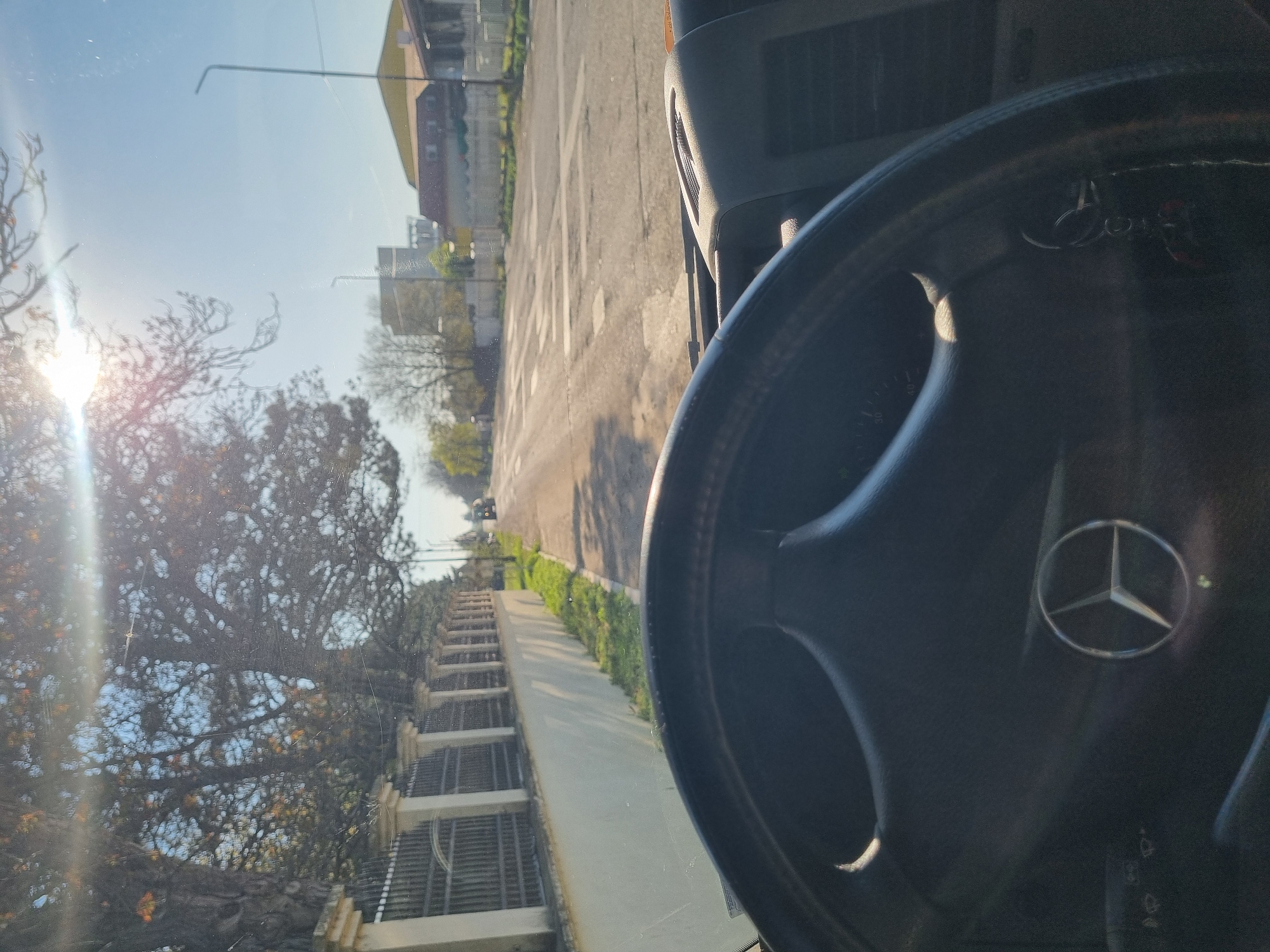 The image is taken from inside a vehicle, showing a Mercedes-Benz steering wheel. Outside the windshield, a street scene unfolds in Ovcharovo, Bulgaria. Buildings are visible along the street, with trees and foliage lining the sidewalk. The street appears to be in a residential area, and the sun is shining brightly.