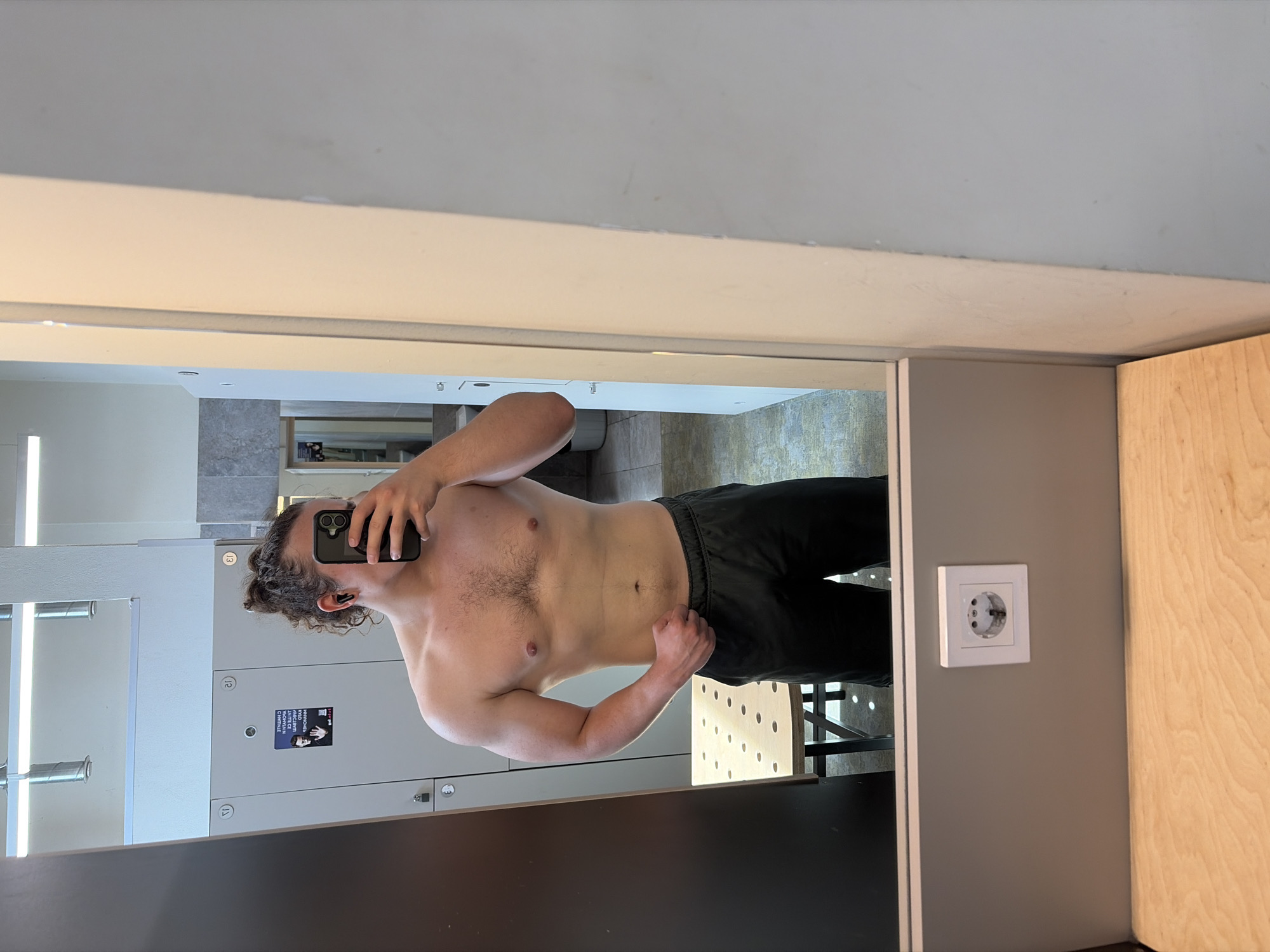 The image shows a person with curly hair, shirtless and wearing black pants, holding a phone in front of their face to take a picture in a mirror. The reflection includes lockers, a light fixture, and what appears to be a bathroom area. Next to the mirror is a grey wall with a power outlet and a wooden surface. The location appears to be a locker room. There are no indications that this photo was taken in the city of Plovdiv, Bulgaria.