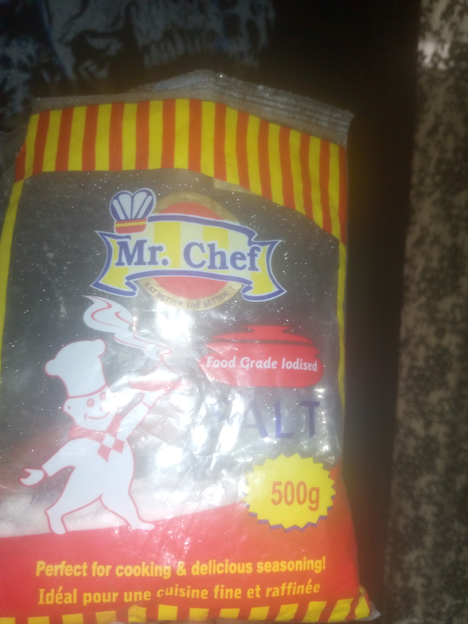 An exterior shot shows a bag of Mr. Chef salt in the city of Unknown, Unknown. The bag is primarily red, yellow, and blue. The Mr. Chef logo, featuring a chef character and a crown, is visible at the top of the bag. Below the logo, text reads "EAT BETTER, LIVE BETTER". Further down, "Food Grade Iodised" is written on a red oval. The word "SALT" is prominently displayed in large blue letters. A yellow starburst with "500g" is to the right of the word "SALT". The bottom of the bag has text in English and French: "Perfect for cooking & delicious seasoning! Idéal pour une cuisine fine et raffinée". The background is dark and out of focus, appearing to be a textured surface, possibly fabric or carpet. The lighting is dim, and the image is somewhat blurry.