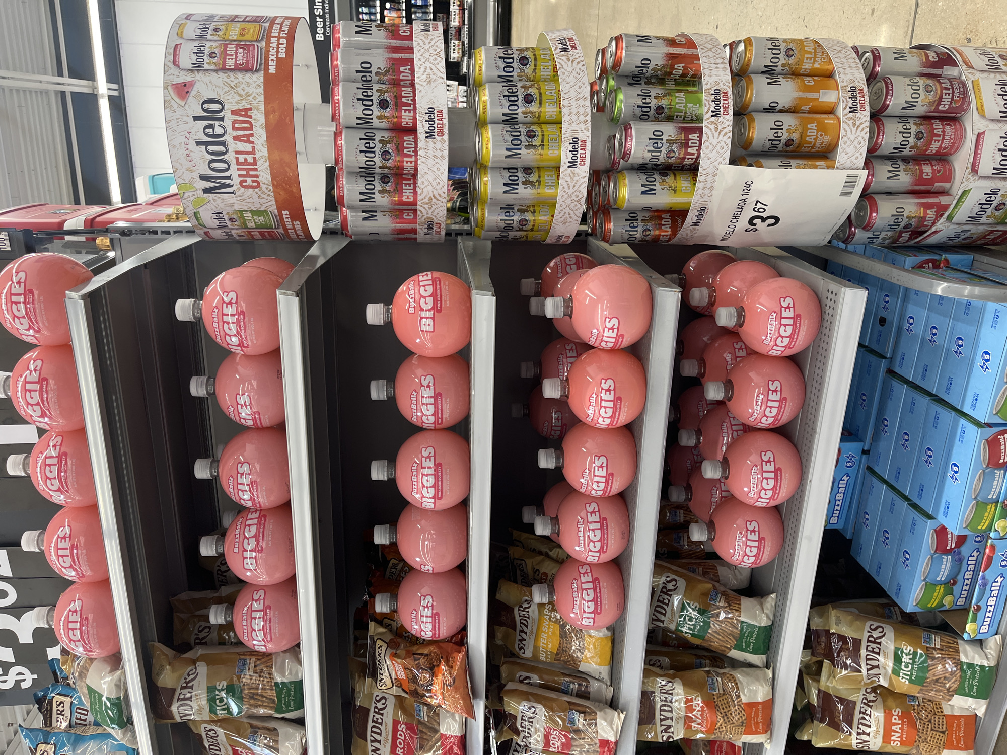 This is a photograph taken inside a grocery store in San Antonio, United States of America. The image shows a retail display shelf stocked with various alcoholic beverages and snack items. Prominently featured are rows of "BuzzBallz BIGGIES" pink lemonade-flavored alcoholic drinks and multiple packs of "Modelo Chelada" beer. Beneath the "BuzzBallz" are bags of "Snyder's" brand pretzels, including sticks, snaps, and rods. The display is set up in a well-lit area with typical grocery store fluorescent lighting visible in the upper part of the image. A price tag for the "Modelo Chelada" beer is clearly displayed, showing a price of $3.67. Boxes of individual "BuzzBall" drinks are also visible.