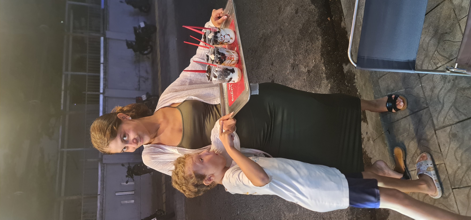 The image captures a woman and a young boy outdoors, likely in the evening or at night, given the artificial lighting and muted colors. The scene is set in an urban environment, specifically in Đà Nẵng, Vietnam, as indicated by the provided context.
The primary subjects are a woman and a child. The woman, with light brown hair, is wearing a dark green tank top with a light-colored, sheer wrap draped over her shoulders. She has a somewhat serious or attentive expression, looking slightly to the side. She is holding a silver tray with four clear plastic cups, each containing a dark-colored drink (possibly bubble tea or a similar beverage with dark toppings like boba or Oreo pieces) and a red spoon/straw. One cup has a penguin design, and another has "MIX" visible on it, while a third partially shows "KOREAN." A red mat with the word "LUONG" printed in white is visible on the tray beneath the cups. The woman is also wearing a dark-colored, form-fitting dress or skirt that extends below her knees, and dark sandals.
The child, a boy with curly light brown hair, is wearing a white short-sleeved t-shirt and dark blue shorts. He is standing close to the woman, looking up at her with an open mouth, as if speaking or reacting to something she is saying or doing. His hands are near his face, with one finger extended.
In the background, there are blurred structures that appear to be buildings and parked motorcycles, consistent with a street or market setting. The ground is paved, showing some texture and what might be damp spots or shadows. A metal chair is partially visible on the right. The overall impression is a casual, public interaction, possibly involving the purchase or distribution of the drinks.