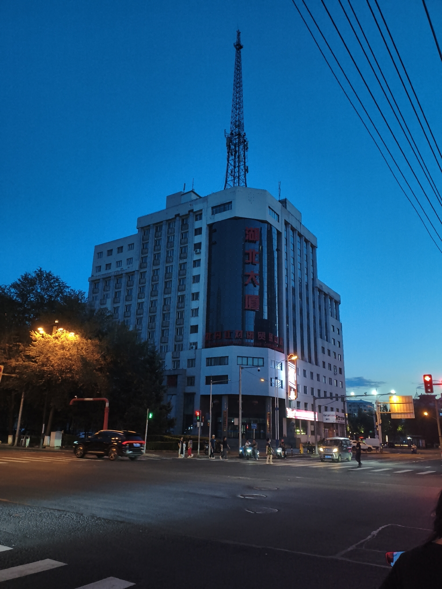 In Dong'an, China, an urban street scene unfolds at dusk. A tall, multi-story building dominates the frame, its facade adorned with vertical rows of windows and illuminated signage in Chinese characters. Atop the building, a lattice-like telecommunications tower pierces the deep blue evening sky. Power lines stretch diagonally across the upper right portion of the image.
The street level features a busy intersection with traffic lights. A dark SUV drives away from the viewer towards the left. Pedestrians are visible crossing the street at a marked crosswalk, illuminated by streetlights and the glow of vehicles. A street lamp casts a warm, orange light on the trees lining the sidewalk to the left of the building. The sky is clear, transitioning from a bright blue to a darker hue as night approaches. The overall impression is of a modern city transitioning from day to night.