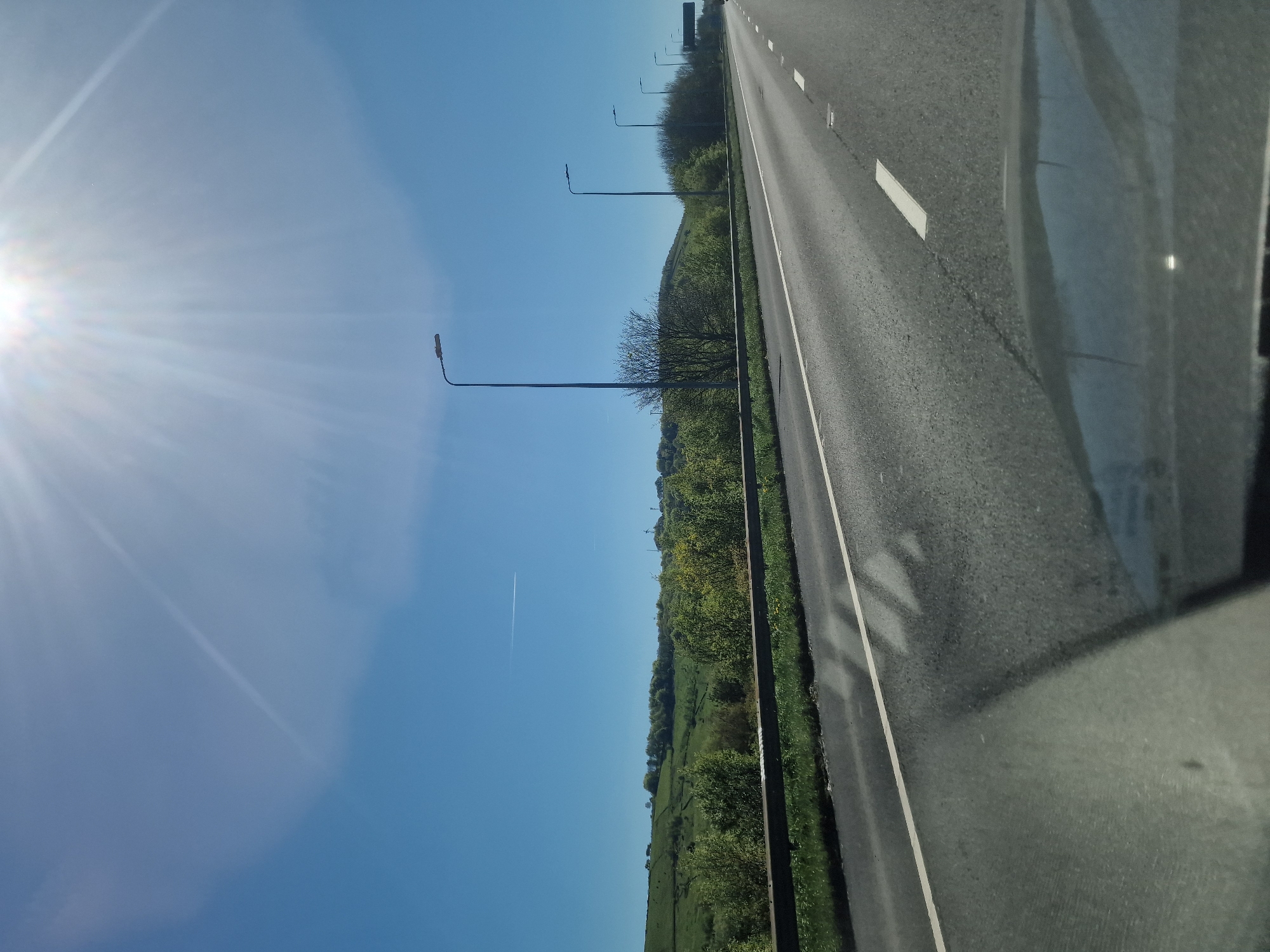 The image depicts a bright, sunny day on a motorway or major highway, likely taken from inside a moving vehicle in Kirklees, United Kingdom. The perspective looks forward and slightly to the left, with the multi-lane road occupying the right side of the frame, receding into the distance. White road markings and reflective studs are visible on the asphalt.
To the left of the road, a narrow grassy verge leads to a series of tall, slender streetlights. Beyond these, a dense line of green trees and bushes forms a barrier, behind which a expansive, undulating landscape of vibrant green fields and rolling hills stretches towards the horizon. The sky is a clear, light blue, dominating the upper portion of the frame. A faint, white contrail from an aircraft is visible high in the sky above the landscape.
The sun is a prominent feature in the upper-left corner, emitting a strong lens flare and bright rays that spread across the sky, highlighting the clear weather conditions. On the far right, the dark interior of the vehicle, possibly a window frame, is visible, along with reflections on the glass showing distorted shapes of the road and surrounding elements. The scene conveys a sense of open-road travel through a picturesque, rural or semi-rural environment under brilliant daylight. No people or legible text are visible.