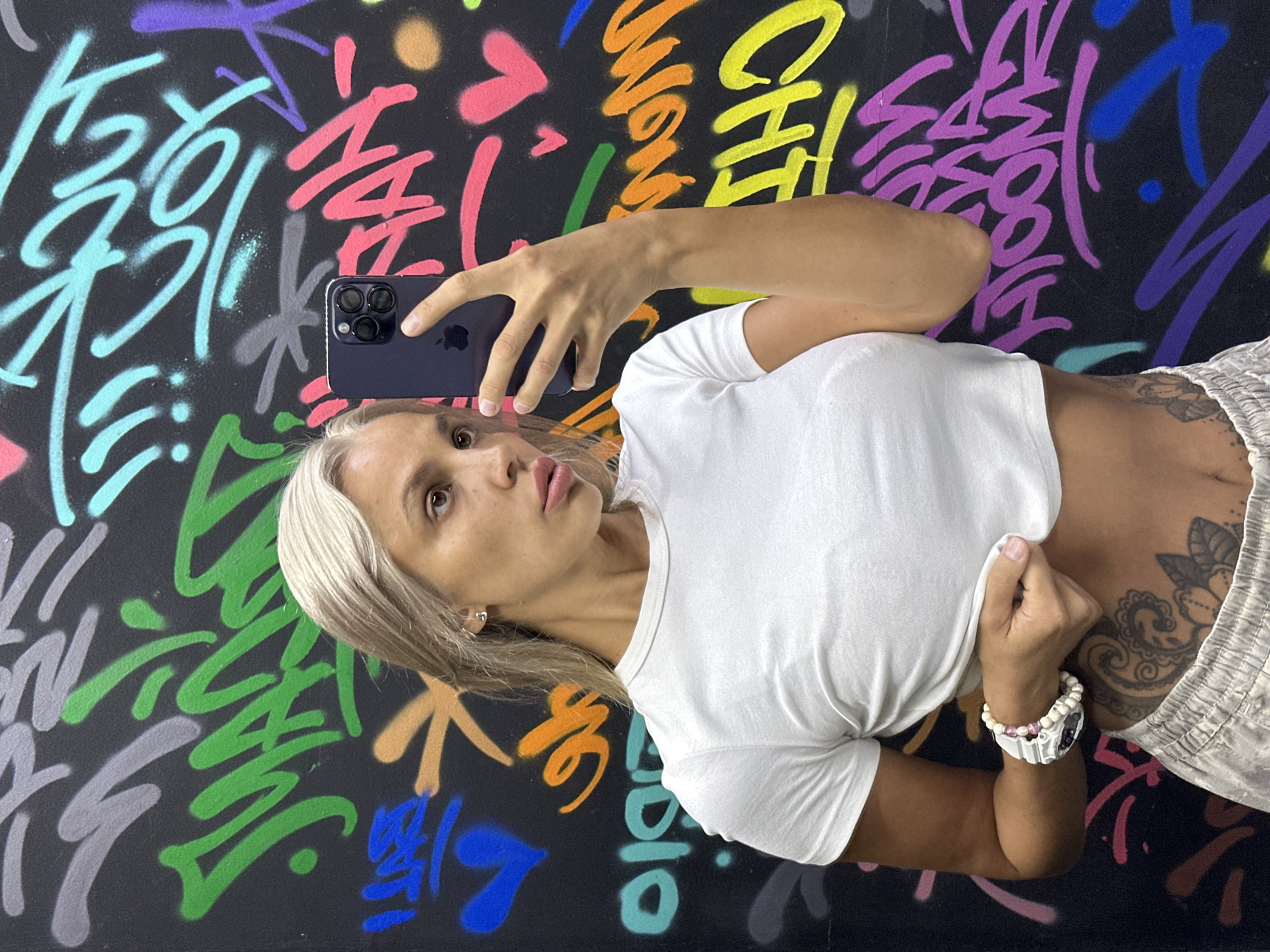 A woman with blonde hair is taking a selfie in front of a graffiti-covered wall. She is wearing a white crop top and light-colored sweatpants. She has a watch on her left wrist and a tattoo on her stomach. The graffiti on the wall is colorful and features various abstract shapes and letters. The location is described as Kuta, Indonesia.