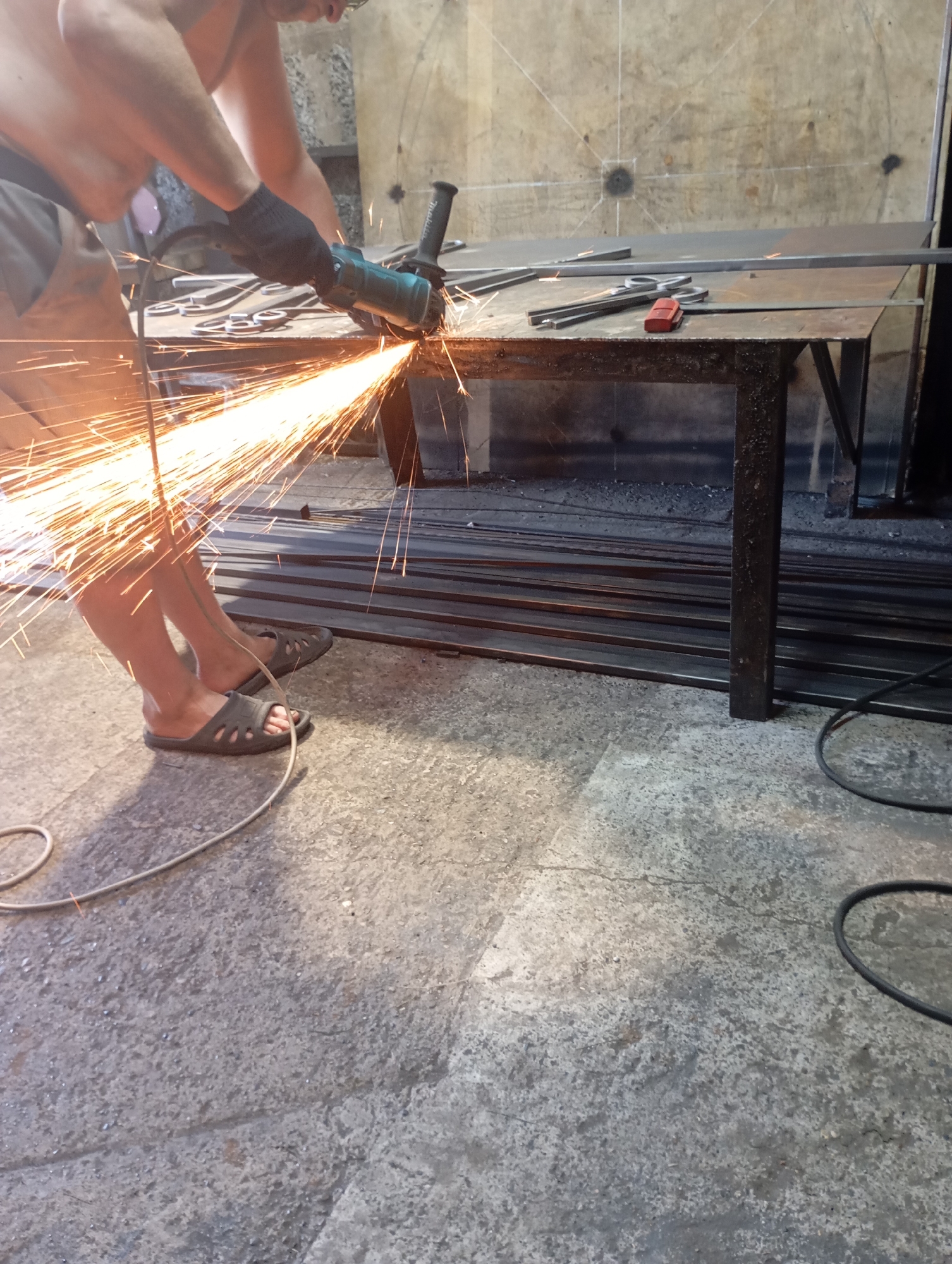 A person is using an angle grinder to cut metal, creating a shower of sparks. They are wearing black gloves and grey shorts. The person is standing in what appears to be a workshop or industrial setting, with metal bars stacked on the floor and a workbench in the background. The floor is concrete, and there are electrical cords visible. The image does not provide any specific visual cues to pinpoint a location within the city of Фоки, Russia.
