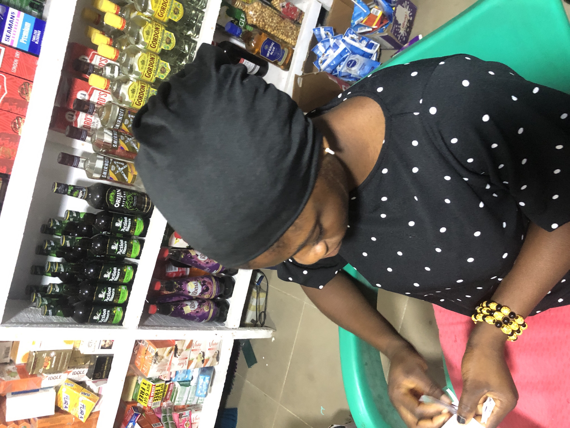 A woman wearing a black dress with white polka dots and a black headscarf is seated in a green chair. She is holding a small white object and a metal tool in her hands, appearing to be engaged in a task. To her left, a pink surface is visible. The background is filled with shelves stocked with various items, including bottles of alcoholic beverages like Gordon's Dry Gin and Smirnoff, as well as packaged goods such as Ribena drinks and other colorful boxes. The setting appears to be a shop or store. The overall lighting suggests an indoor environment.