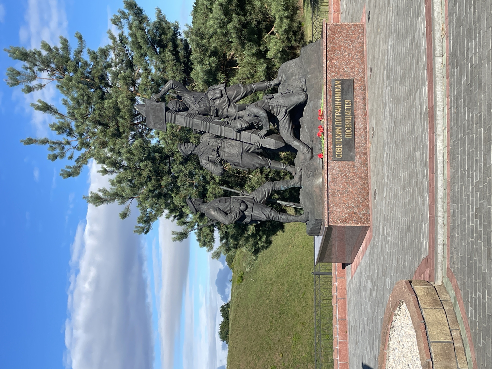 The image shows a monument to Soviet border guards located in the city of Brest, Belarus. The monument is a bronze sculpture of four soldiers, three standing and one kneeling, with one of the standing soldiers holding a border post above his head. Red flowers are placed at the base of the monument, which has a granite pedestal with an inscription in Russian. Behind the monument is a large evergreen tree, and to the left is a grassy hill. The ground in front of the monument is paved with interlocking bricks, and there is a low brick wall and metal railing surrounding the monument area. The sky is blue with scattered clouds.