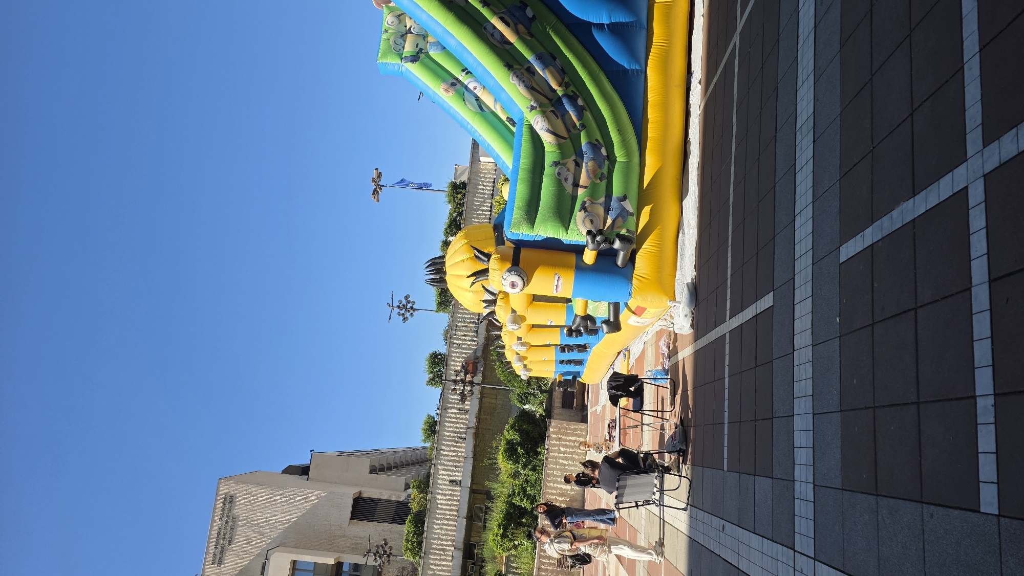 The image shows an outdoor scene in Blagoevgrad, Bulgaria. In the foreground, there is a large inflatable bounce house featuring Minion characters in shades of yellow, blue, and green.
To the right, there is a paved area of brick and concrete squares. Further back, chairs and people are present, appearing to be related to the bounce house activity. The people are adults.
Behind the bounce house, there is a building with dense green vegetation climbing over the walls. The sky is bright blue with no clouds visible. Street lights are visible on the other side of the inflatable.