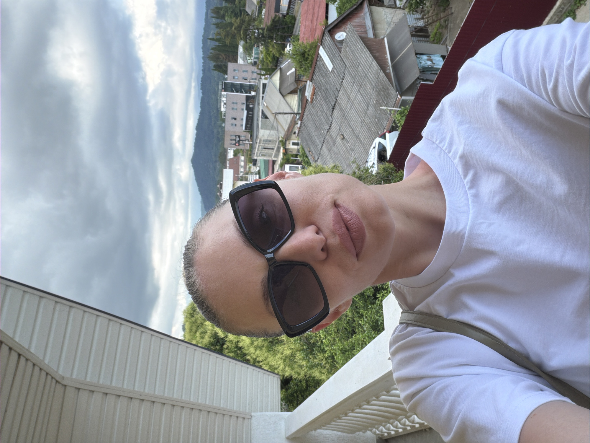 A woman with blonde hair pulled back and wearing large dark sunglasses takes a selfie on a balcony in Sochi, Russia. She is wearing a white t-shirt and a light green strap across her chest. Behind her, a cloudy sky looms over a town with numerous buildings and trees, and in the distance, mountains are visible.