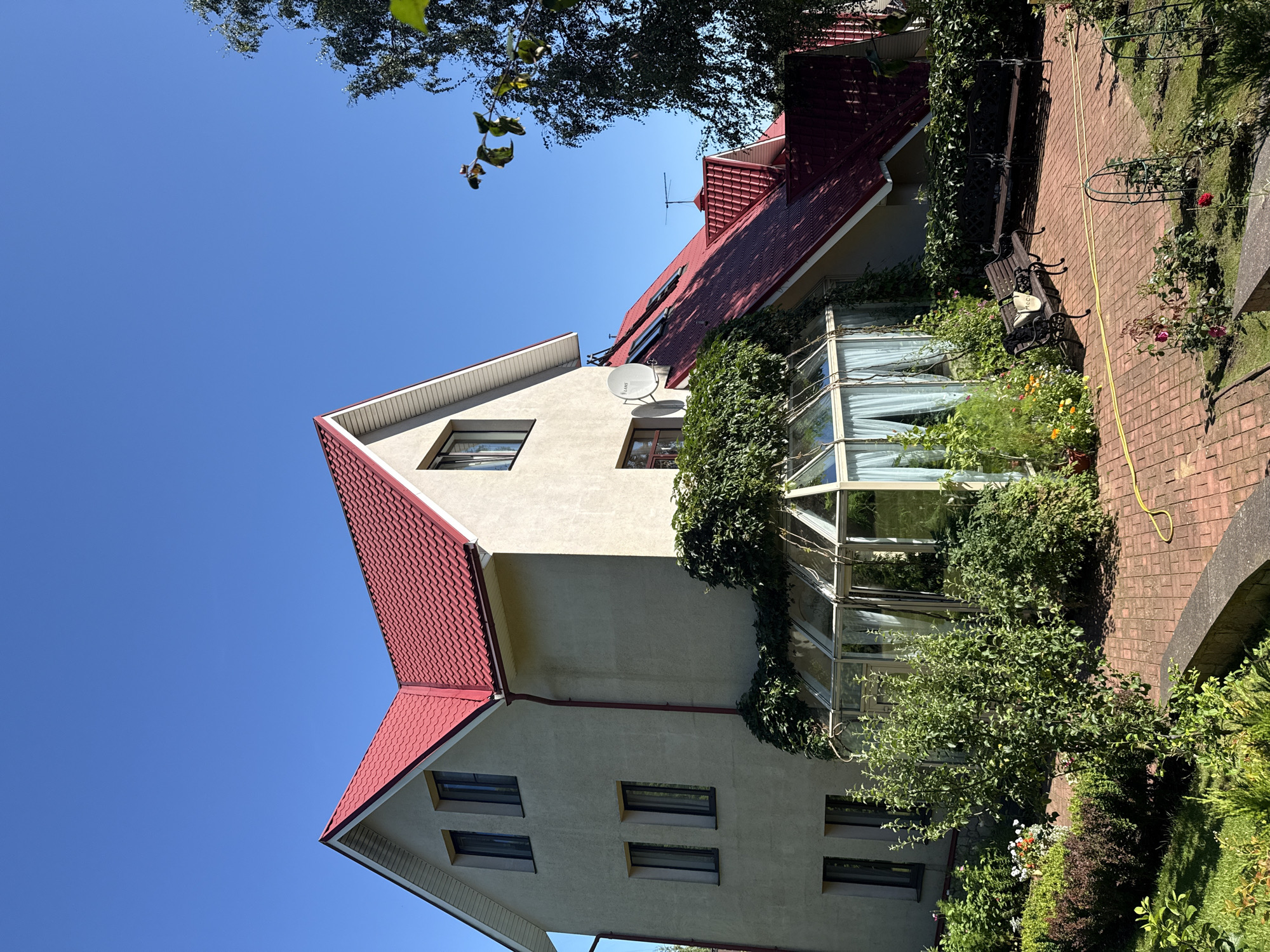 A brightly lit, eye-level, long shot captures a large, light-colored, multi-story house with a vibrant red metal tile roof under a clear blue sky in Yukki, Russia. The house features multiple windows and a prominent glass conservatory attached to its side, draped with light-colored curtains. The conservatory is partially covered by ivy. The surrounding yard is well-maintained, with a brick patio, lush green lawns, flower beds with blooming flowers, and various trees and bushes. A wooden bench with a white bag resting on it, and another dark metal bench, are visible on the patio. A yellow garden hose lies coiled on the brickwork. The overall scene suggests a residential property in good condition on a sunny day.