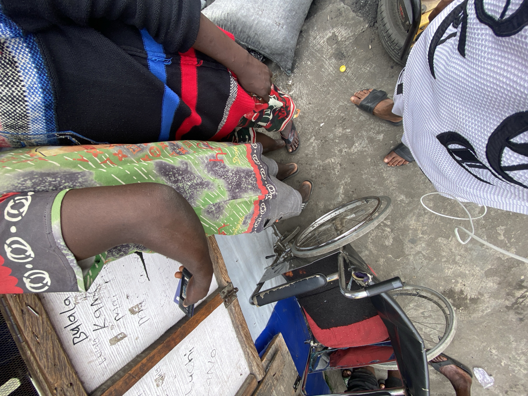 The image depicts a scene in Lagos, Nigeria. It shows several people, including someone sitting on a chair with a blue and red striped blanket. Another person is using a wheelchair with a green and gray patterned cloth covering their legs. They are using a mobile phone and facing a white board with writing on it. A third person is standing wearing a white and black patterned garment. The environment is a street with a concrete surface. There is a bicycle wheel, a gray bag, and tires visible. The writing on the board includes the word "Bulala".