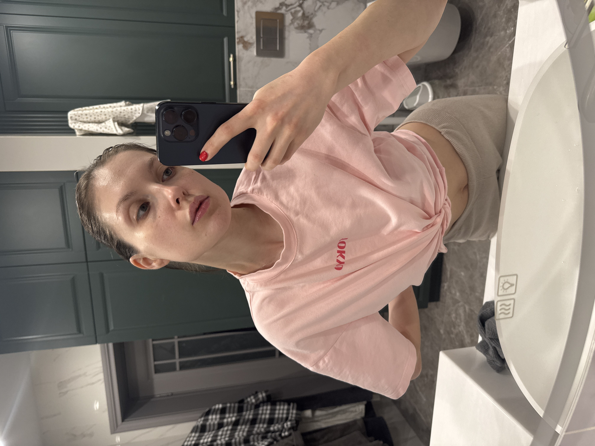 A woman is captured taking a mirror selfie in a bathroom, with the image rotated 90 degrees counter-clockwise. She holds a dark smartphone, likely an iPhone, in her left hand, which has red-painted fingernails. Her head is slightly tilted, and she looks directly at the camera with a neutral expression, revealing some freckles on her nose and cheeks. Her dark hair appears pulled back and slightly damp.
She is wearing a light pink short-sleeved t-shirt with the word "OKAY" written in red on the front. The t-shirt is tied in a knot at the bottom, exposing a small portion of her midriff, and she wears light grey or beige sweatpants.
The background of the bathroom features dark green, paneled wooden cabinets. Marble-patterned tiles in grey and white are visible on the walls, along with a grey electrical outlet or light switch. A white sink or vanity counter is present on the right side of the frame, with a grey washcloth resting on it. On the mirror's surface, two illuminated touch icons are visible: a light bulb symbol, suggesting mirror lighting, and a symbol with wavy lines, possibly indicating a defogging or heating function. A white cloth with a star pattern is draped over a shelf or object in the upper background, and a plaid shirt is visible on a surface in the lower left corner.