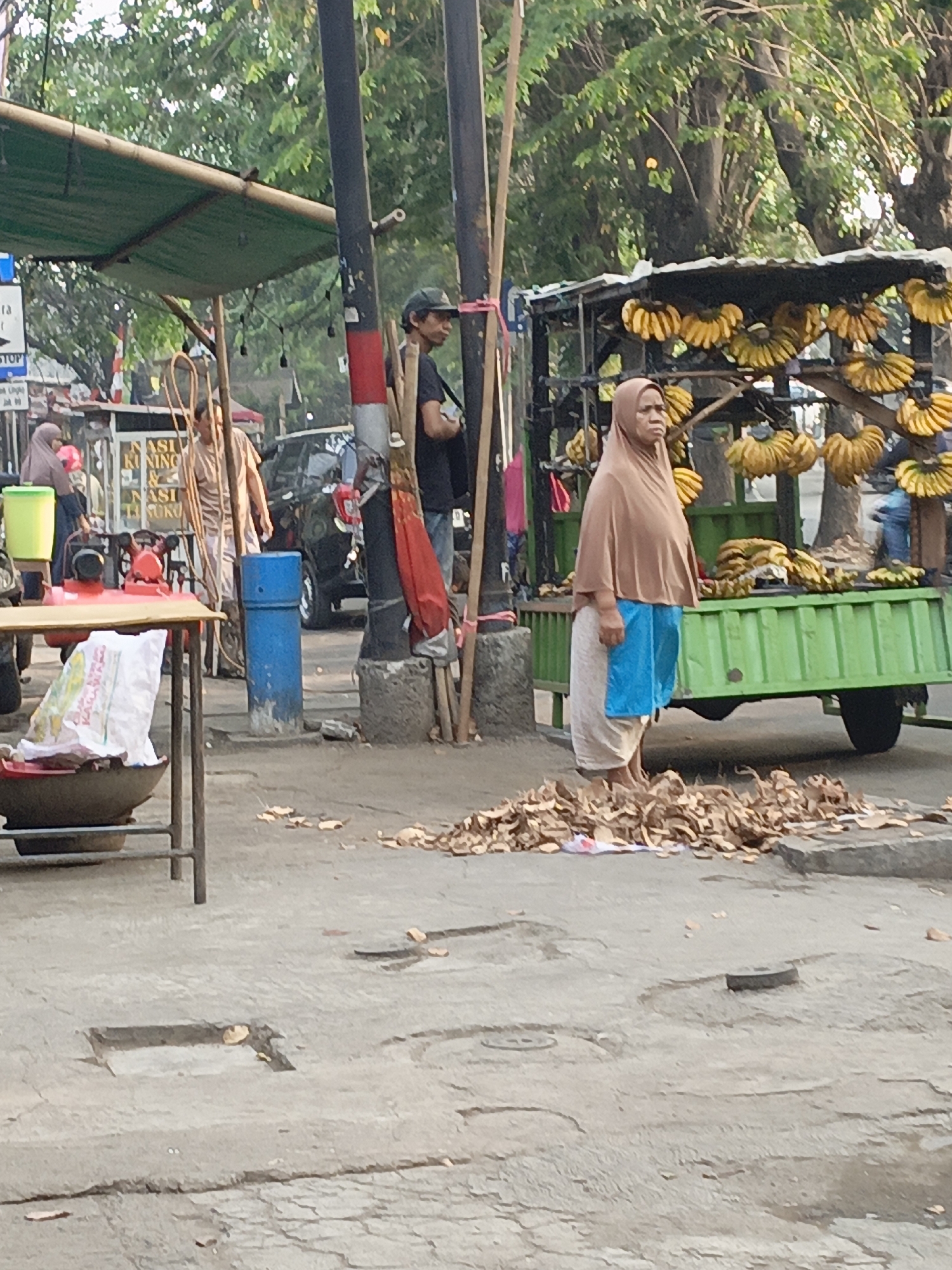 The image shows a street scene in East Jakarta, Indonesia, with a focus on a banana seller. A woman in a brown hijab and blue skirt stands in front of a cart laden with bunches of ripe bananas. The cart is green and has a metal frame. In the foreground, a pile of banana peels is scattered on the ground. To the left, a vendor stall with a green awning is visible, with a person in the background wearing a light-colored patterned shirt. A red machine and a yellow container are on a table at the stall. A blue barrel stands near a lamppost with a red and white striped band. A black car is parked in the background, and trees line the street. The ground is paved with concrete and shows signs of wear and tear, with scattered leaves and debris.