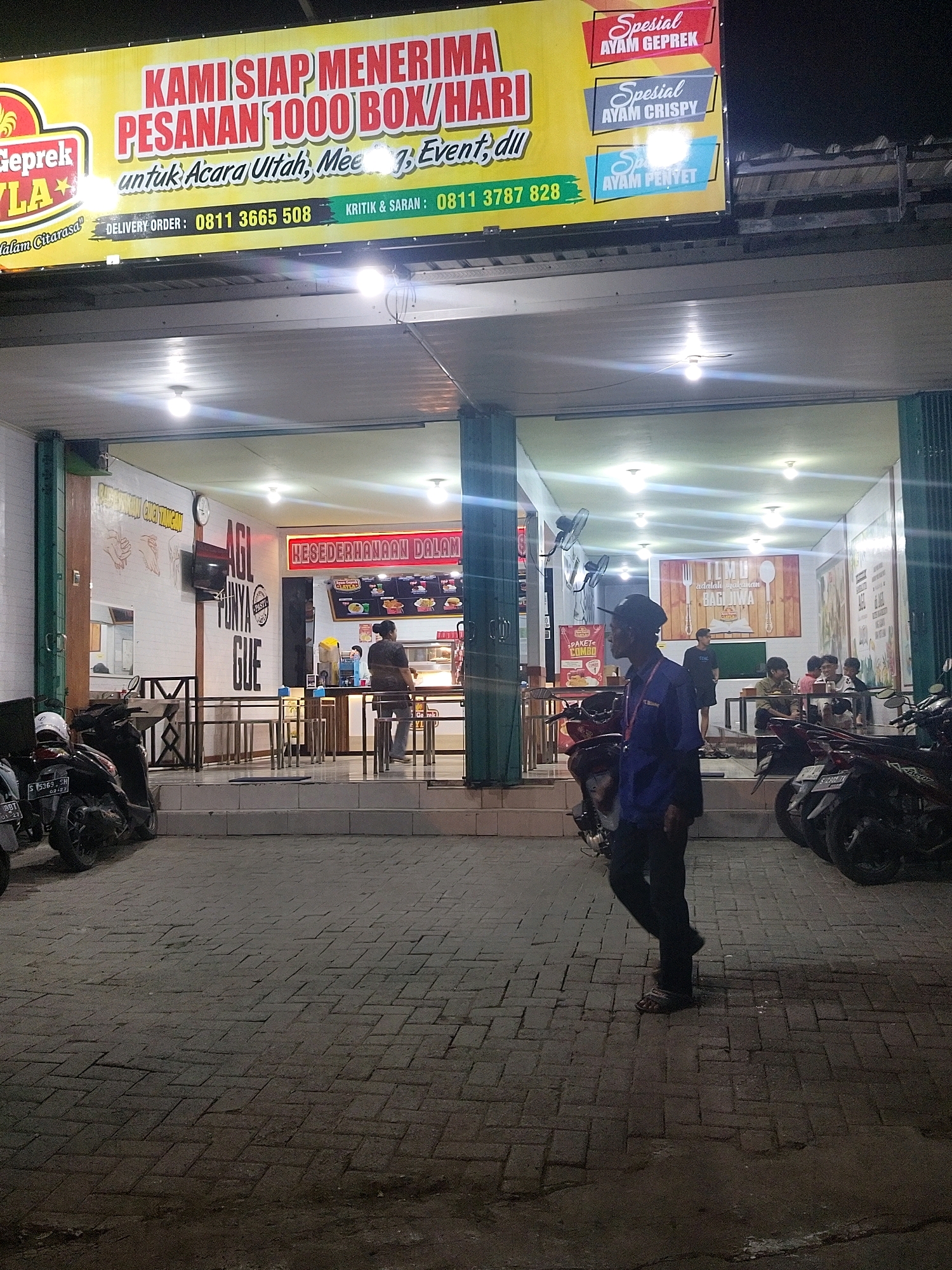 The image shows a storefront at night in what appears to be a commercial area of Mojokerto, Indonesia. A large, illuminated yellow banner above the storefront reads "KAMI SIAP MENERIMA PESANAN 1000 BOX/HARI" (We are ready to accept orders of 1000 boxes/day), along with information about delivery orders and criticism/suggestions. Below the banner, the building has an open facade revealing a dining area with tables and chairs, and staff members behind a counter. Inside, the walls are decorated with themed artwork and a menu board. Outside the storefront, several motorcycles are parked on the paved ground. A man in a blue uniform and dark pants is walking away from the camera in the foreground. The overall scene depicts a food establishment operating in the evening.