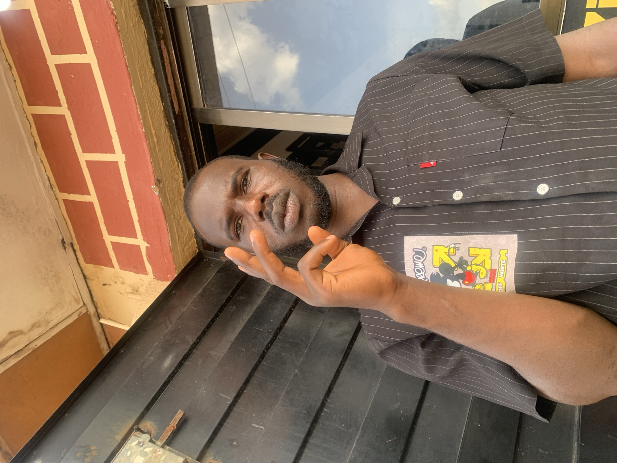 A man with dark skin and a beard is positioned at an angle in the frame. He is wearing a dark, striped shirt with buttons. His hand is raised, with the index finger and thumb touching. Behind the man, there is a window reflecting the sky and clouds. To the left, a brick wall can be seen. The scene appears to be an outdoor setting. No definitive details in the image confirm the location in Jos, Nigeria.