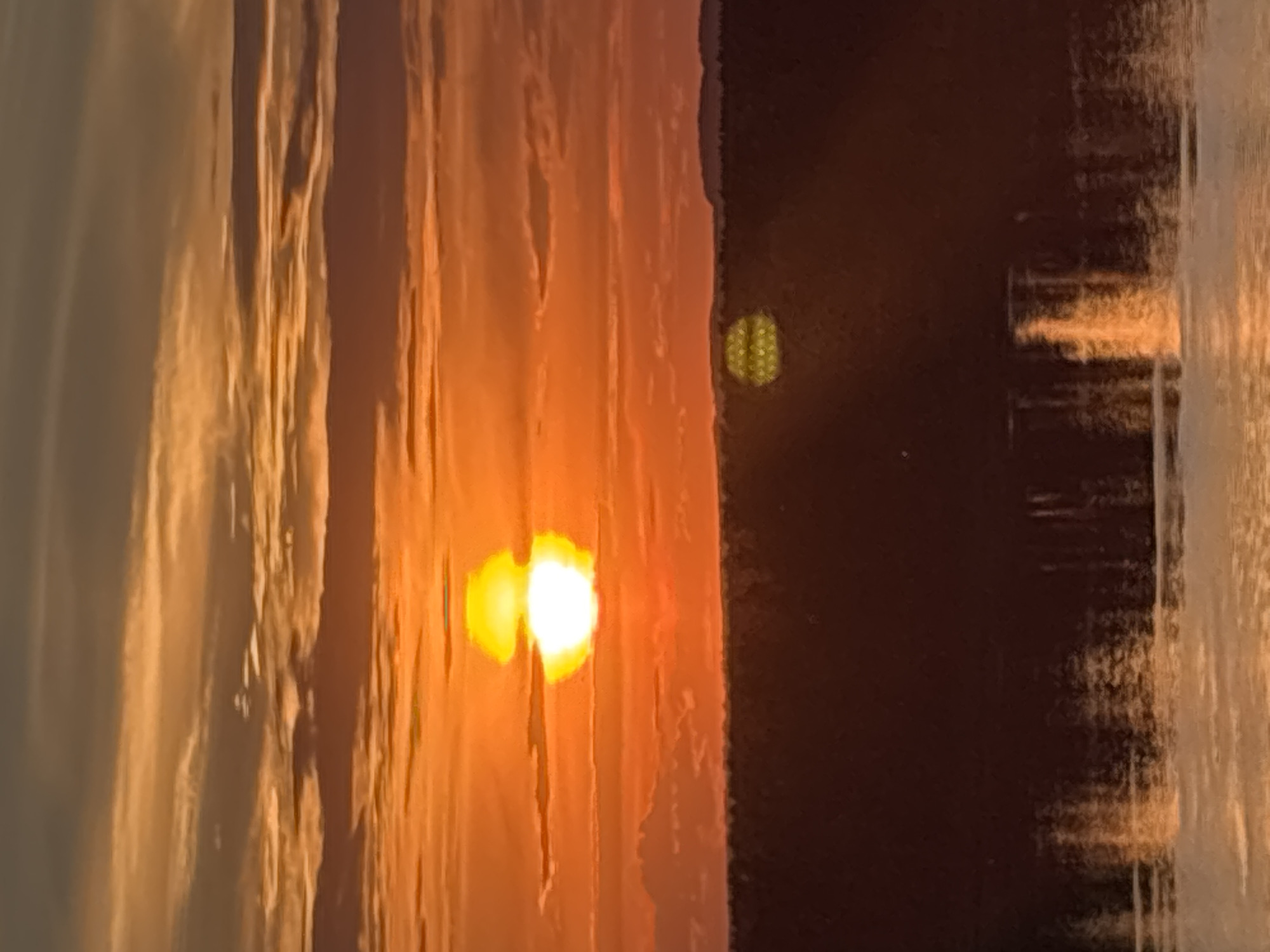 This media file depicts a sunset over a body of water. The sun, a bright orb of light, is partially obscured by textured vertical elements that appear to be clouds or possibly a structure. The colors are dominated by warm oranges and yellows, indicating it is either sunrise or sunset. The right side of the image shows a darker expanse, likely land or a deeper part of the water, with a reflection of the bright sky and sun. There are faint, pixelated shapes visible in the darker area, one of which resembles a honeycomb pattern. The overall mood is serene and picturesque, capturing the natural beauty of the moment. The location context suggests this scene is in Olkhovka, Russia. There is no visible text in the image.