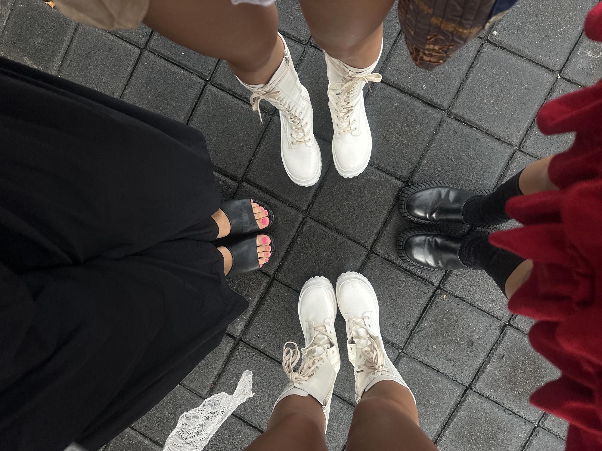 Five people are standing in a circle on a grey tiled surface. One person is wearing black sandals with pink painted toenails and a long black dress. Another person is wearing tall white lace-up boots. A third person is wearing black boots with black socks and a red dress or skirt. A fourth person is wearing white lace-up boots with tan skin. The location is likely in North Kuta, Indonesia, given the prompt.