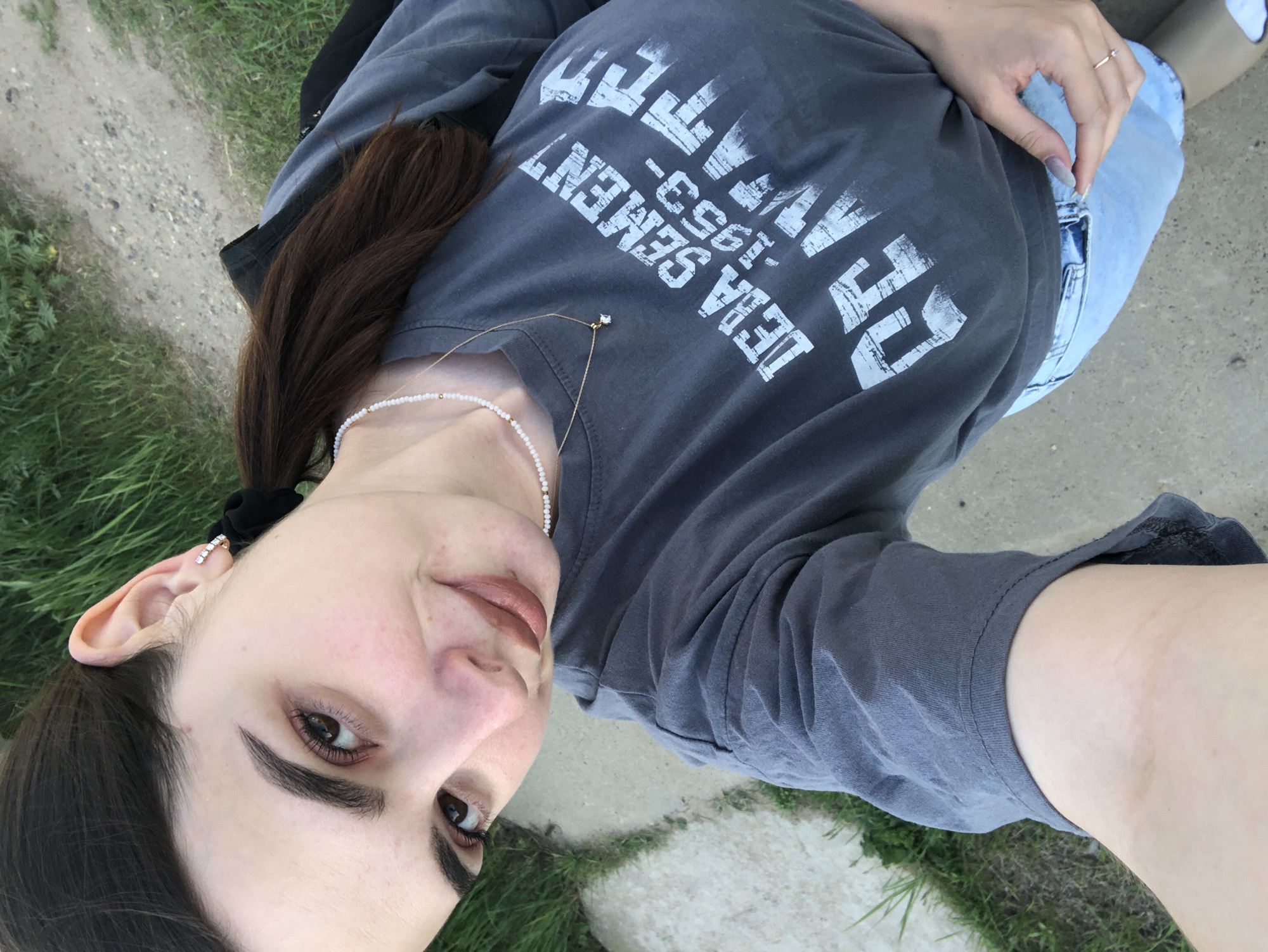A medium-shot, high-angle photograph depicts a woman outdoors, likely in a park or grassy area. She is wearing a grey t-shirt with white lettering that appears to read "Deira Cement - 1953". She also has on light-wash jeans, a pearl necklace, and a gold pendant necklace. Her hair is dark brown, pulled back in a low ponytail secured with a black scrunchie. The woman is looking at the camera. The background consists of a patch of green grass and a concrete path or sidewalk. There is no information available to determine if the location is in Noviy, Russia.