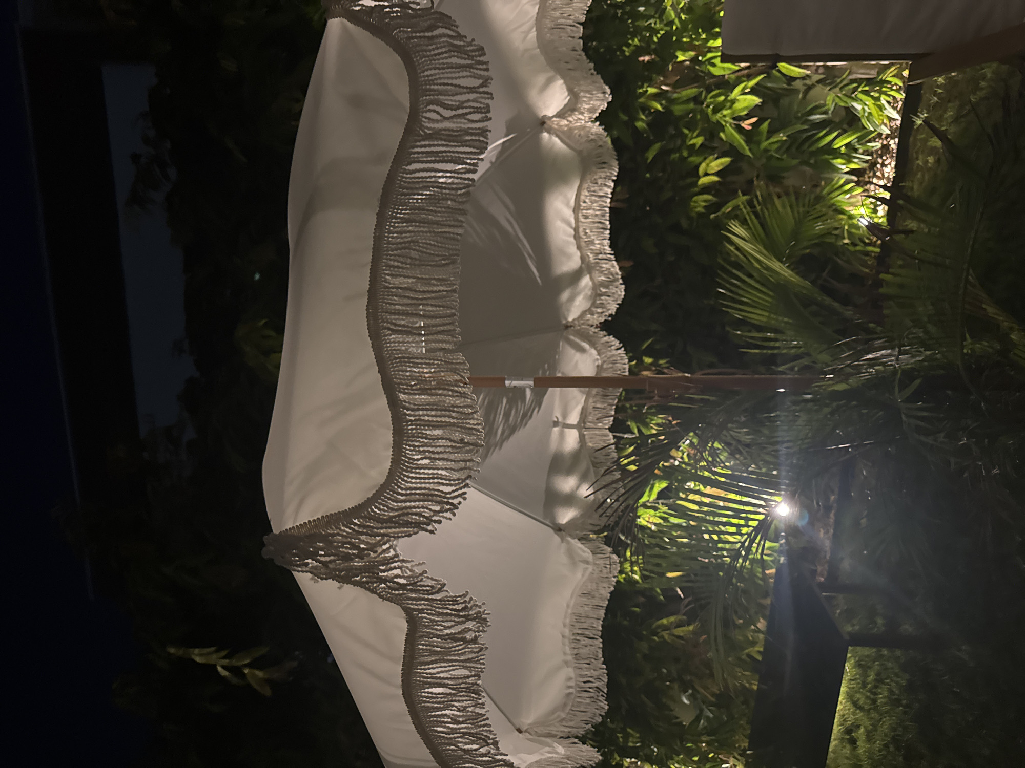 The image captures an outdoor scene at night, possibly in the city of Agios Tychona Community, Cyprus. The focal point is a white patio umbrella with decorative fringe. The umbrella is partially open, revealing its structure and the fabric's texture. Surrounding the umbrella is lush greenery, with some plants illuminated by ambient light, casting shadows and highlights. The background suggests a dark sky with some faint illumination from what could be distant lights or structures. The overall impression is one of a serene, possibly residential or hospitality setting at dusk or night.
